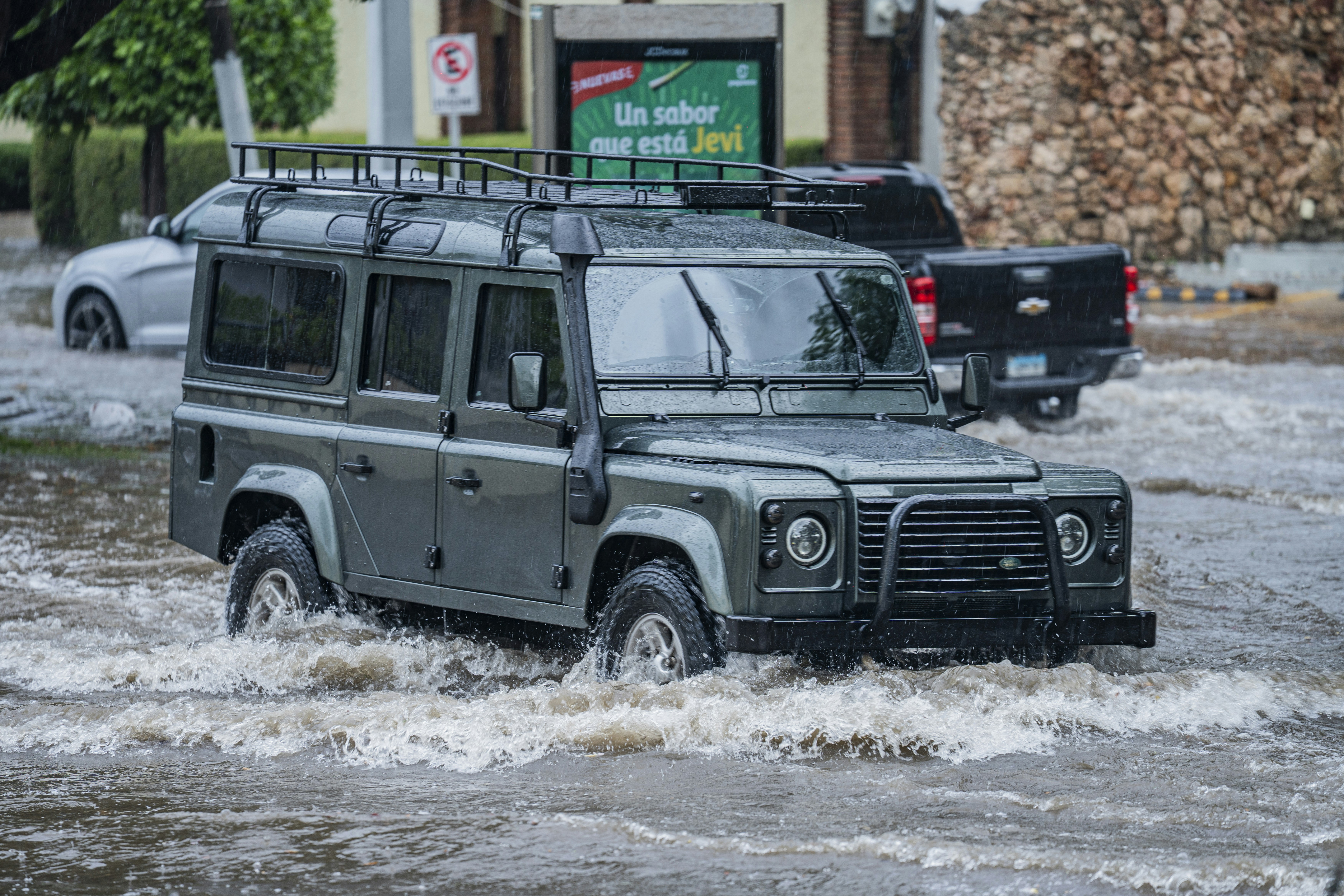 A dark green Land Rover Defender driving through a flooded street during rain