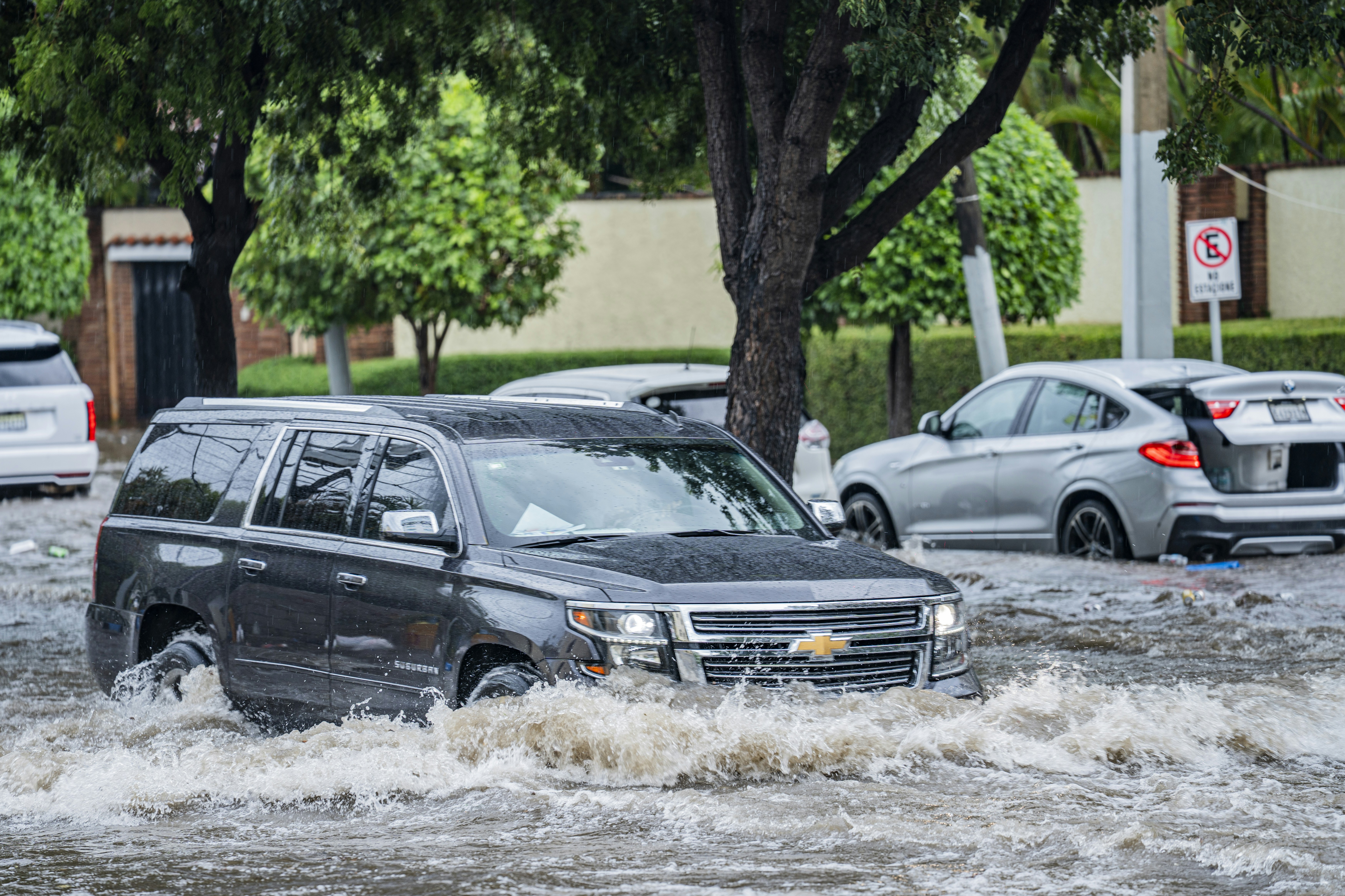 A dark SUV driving through deep floodwater on a street with trees