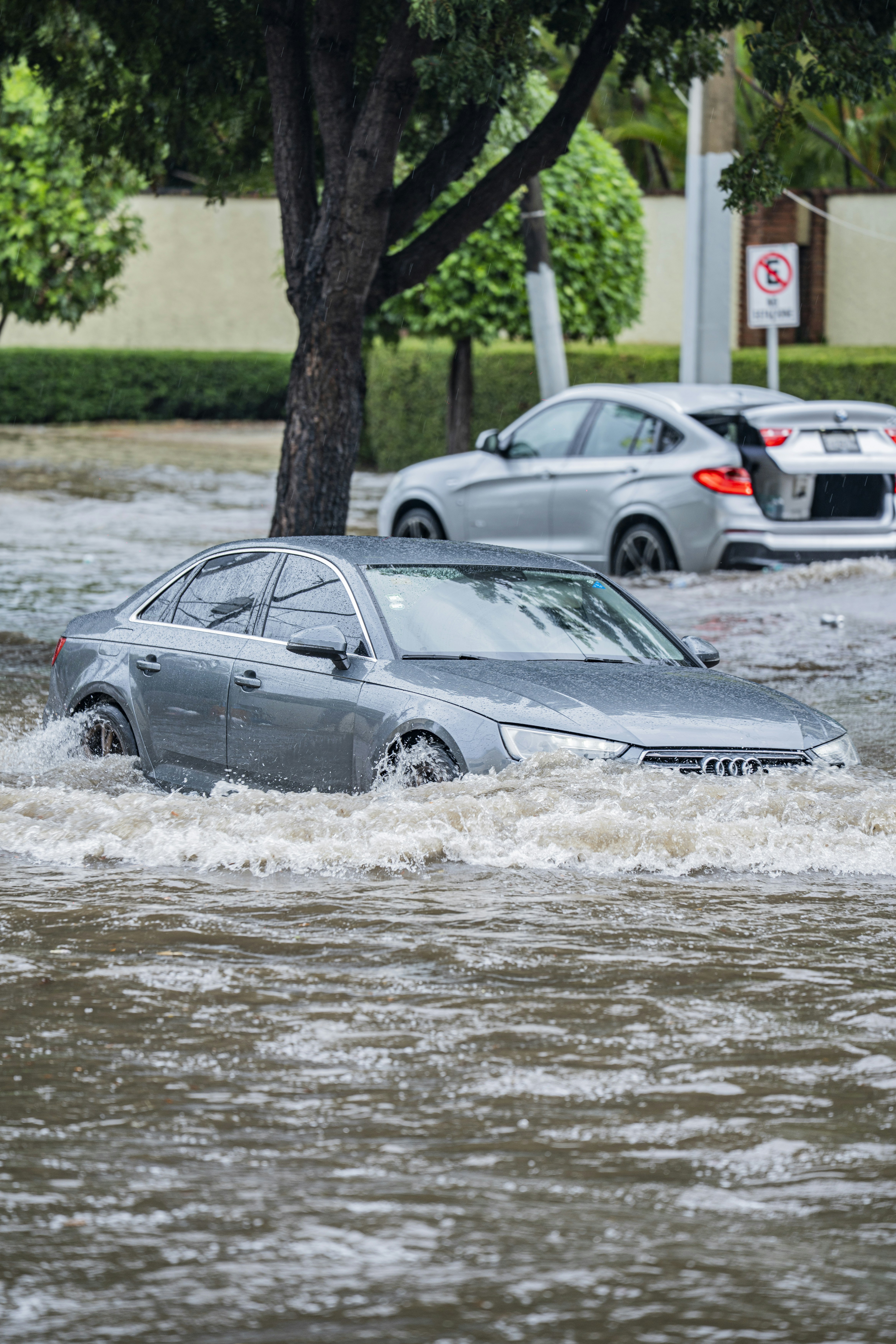 A grey sedan driving through deep floodwater on a rain-slicked city street