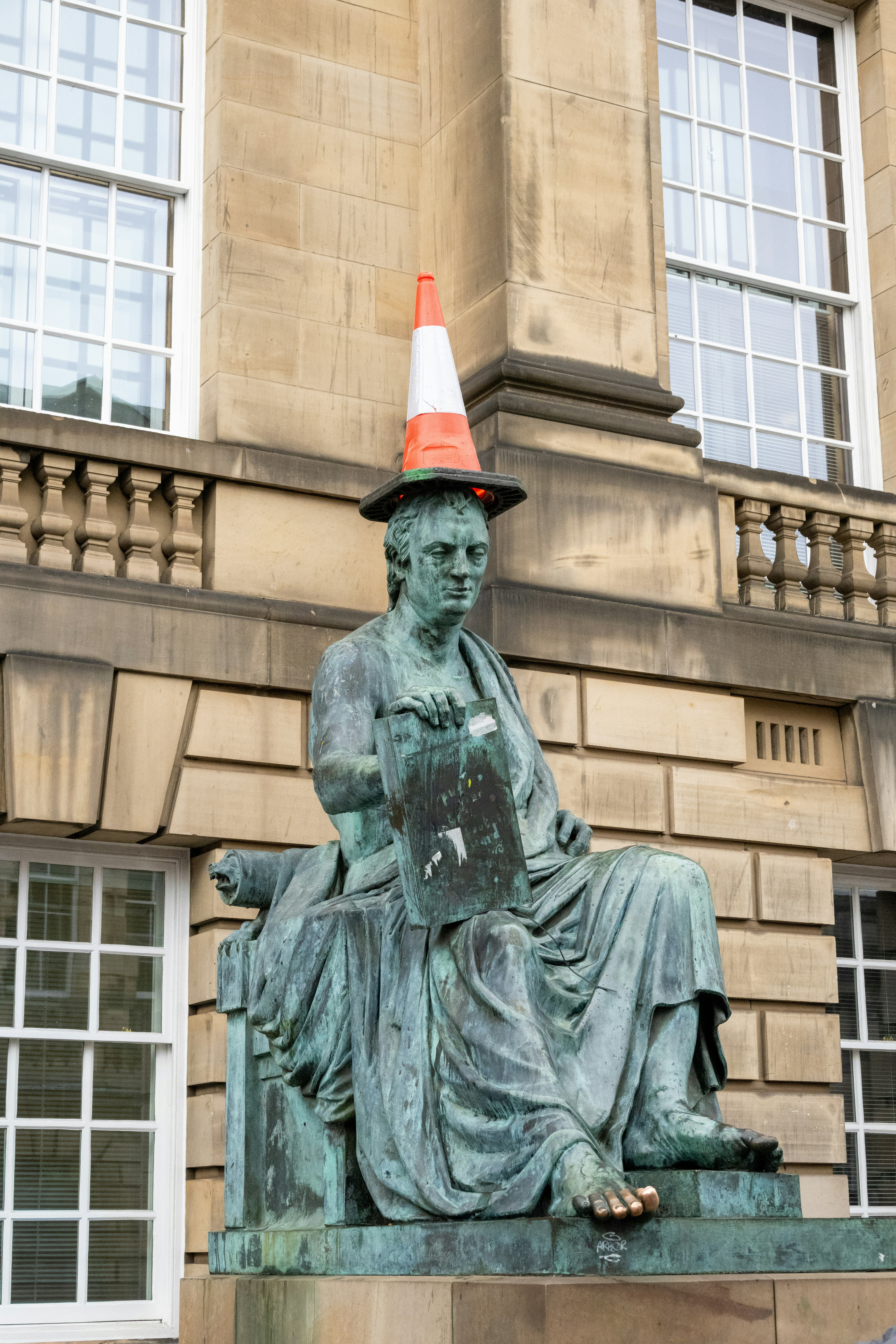 A bronze statue of a seated man wearing an orange traffic cone on his head