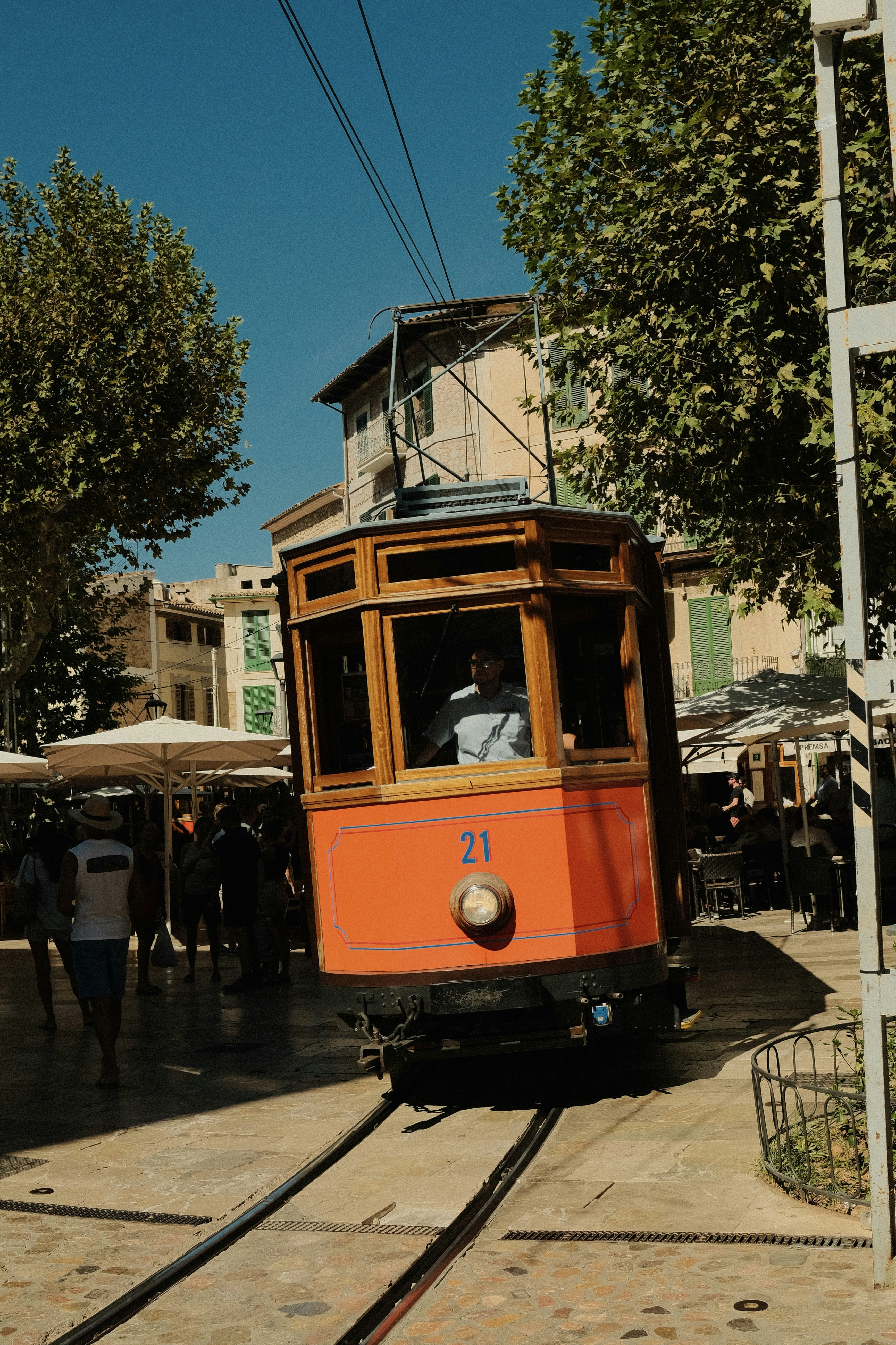 A vintage orange wooden tram moving through a sunlit town square with pedestrians