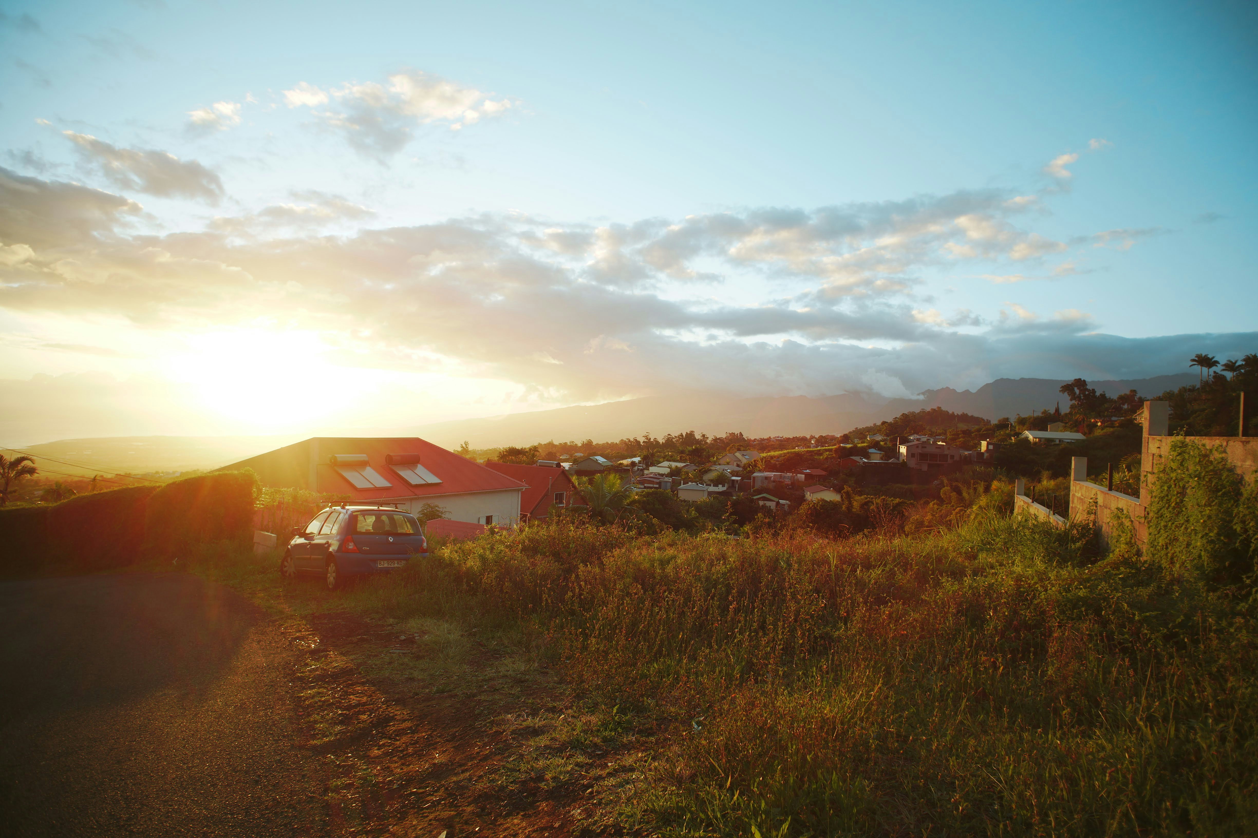 A blue car parked near a house with a red roof at low sun