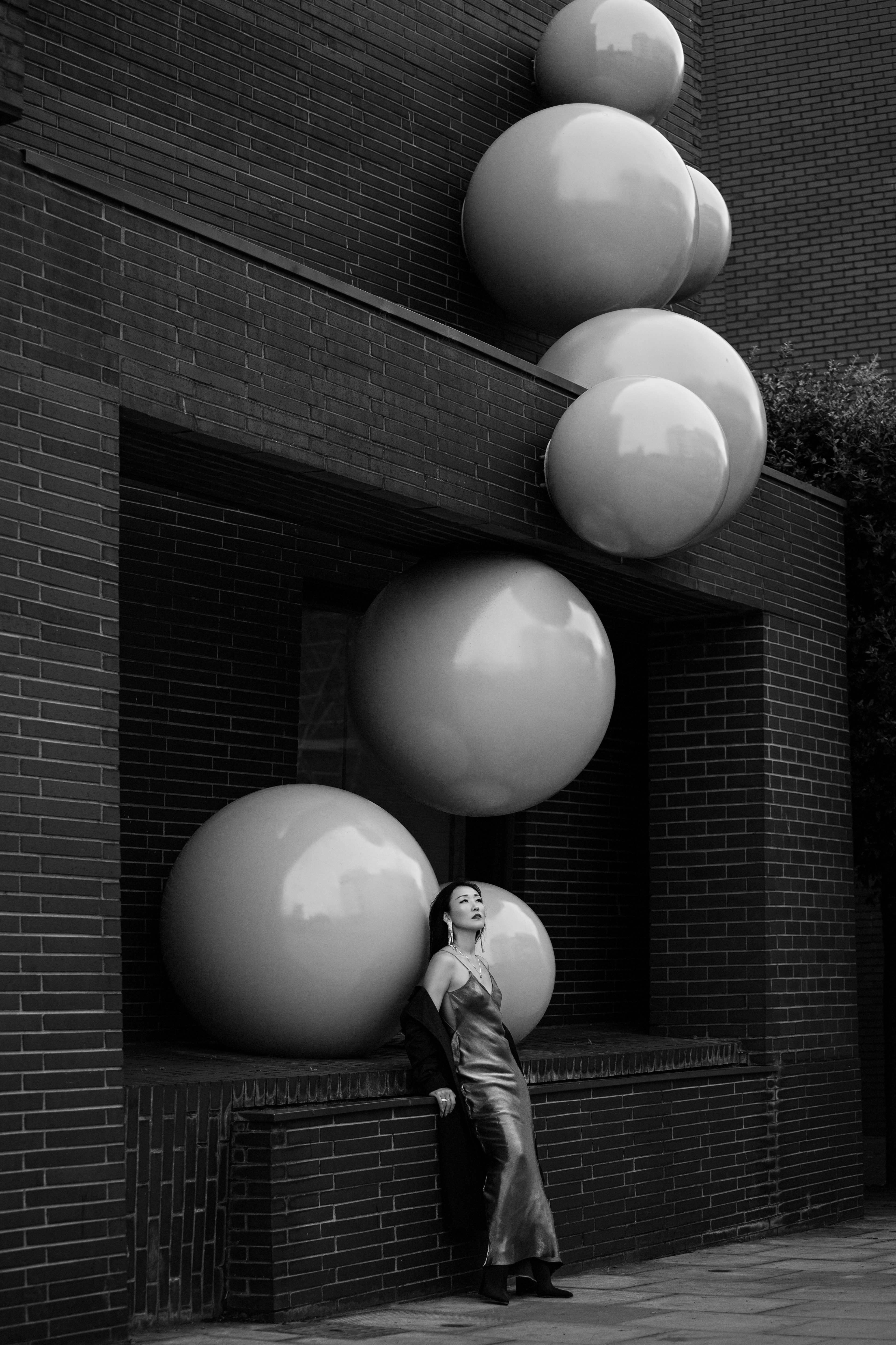 A woman in a slip dress leaning against a brick wall with large spheres