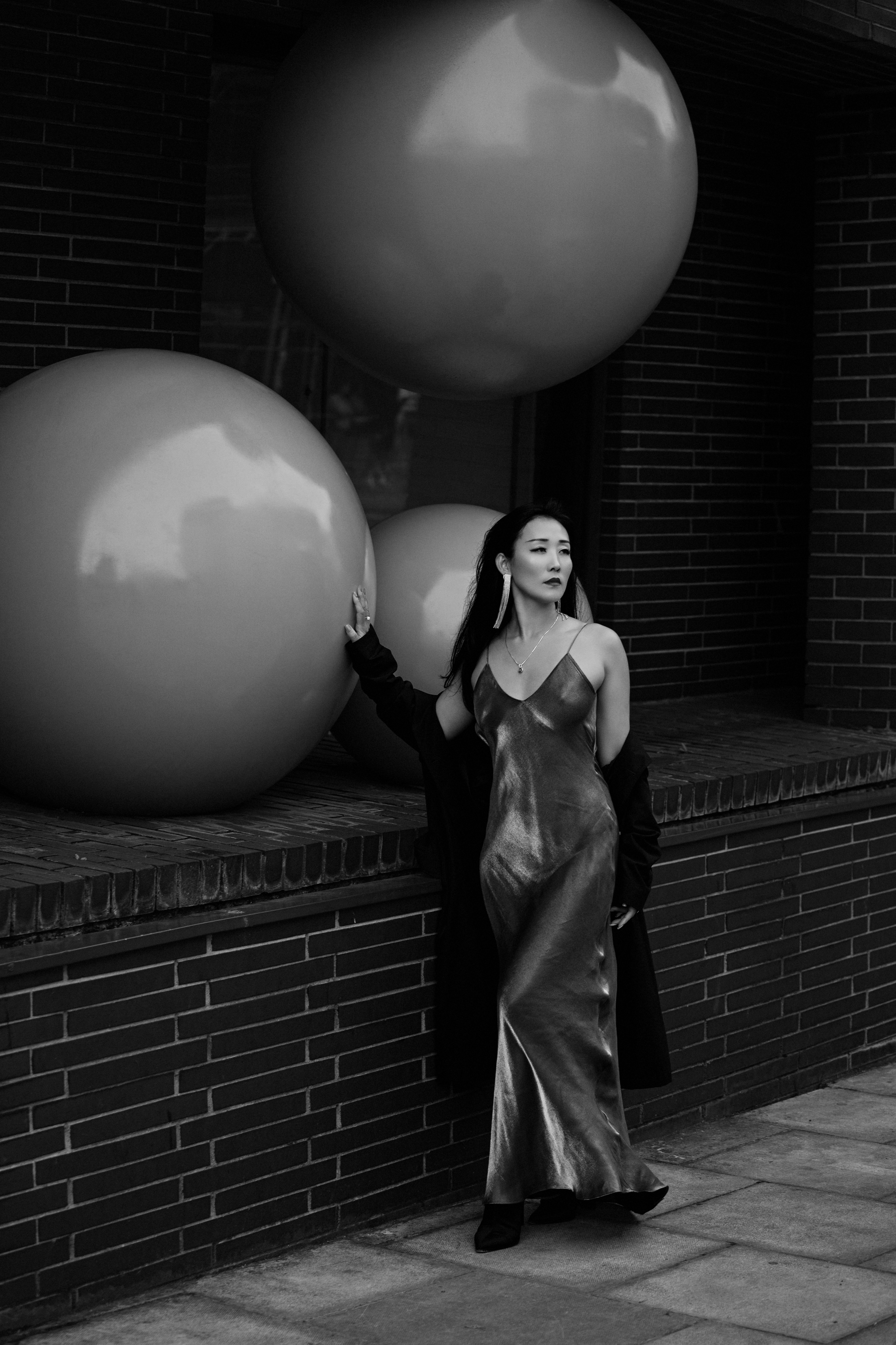 A woman in a metallic slip dress leaning against large spheres on a brick ledge