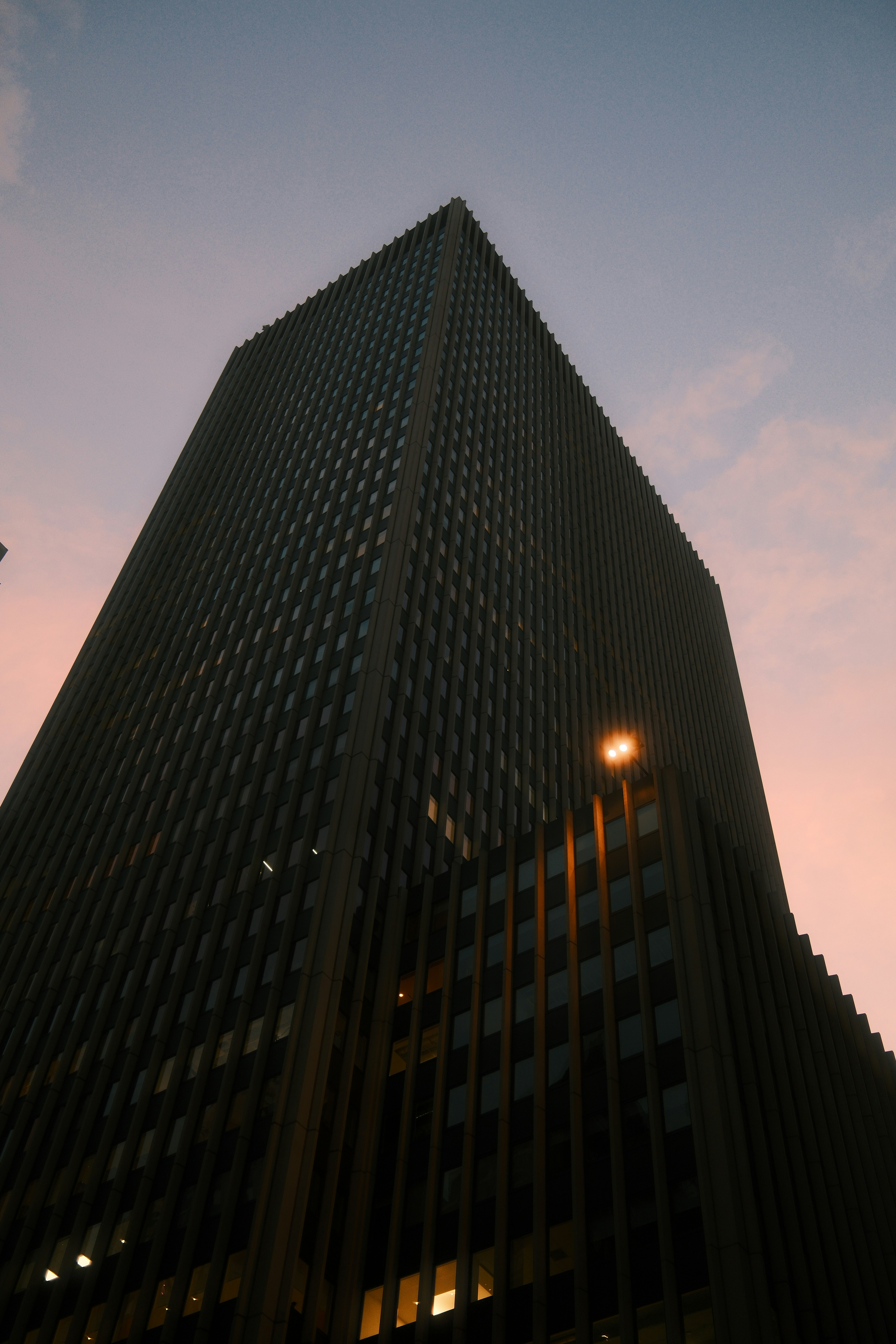 A dark skyscraper against a soft pink sky with a single glowing light