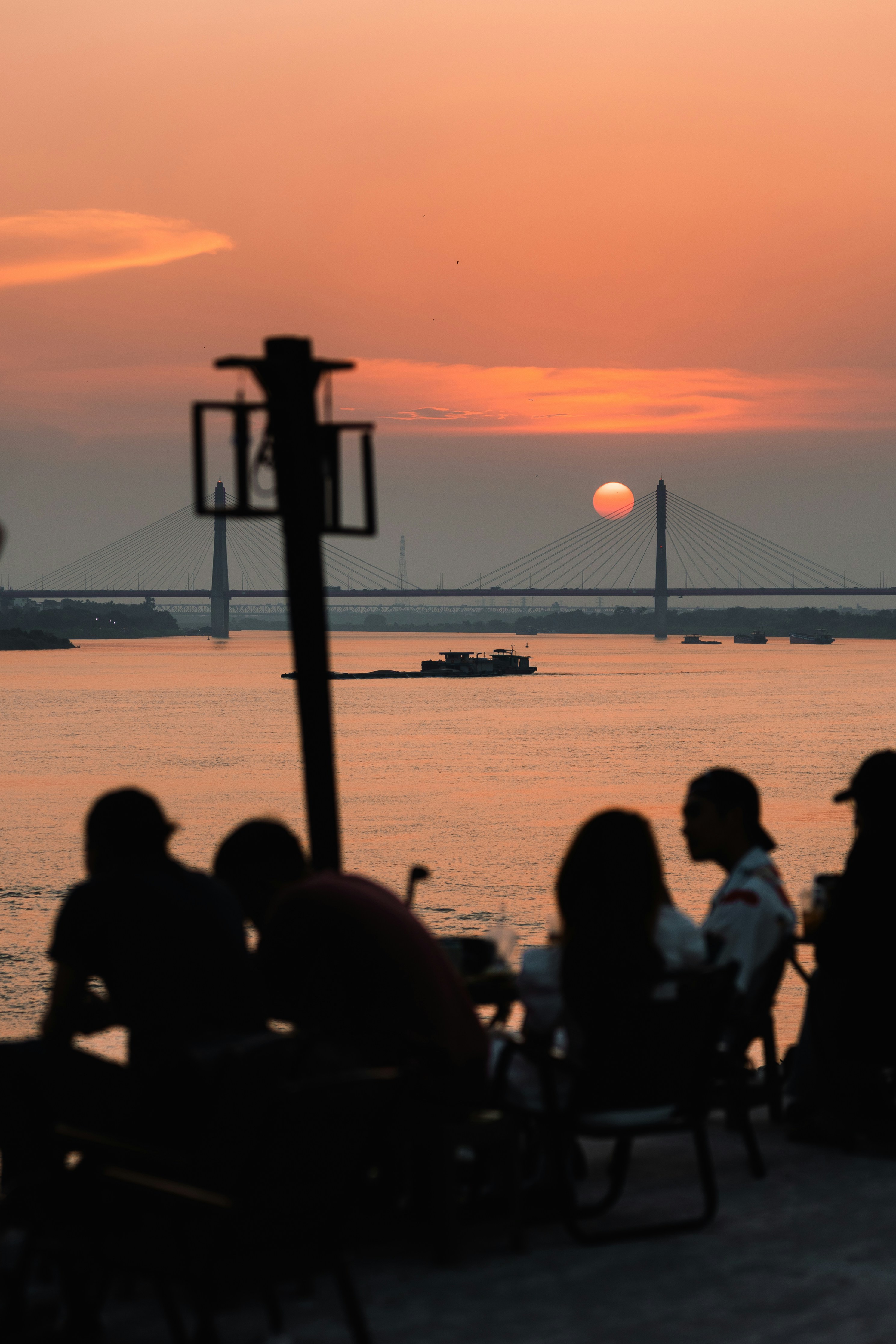 Silhouettes of people sitting at tables overlooking a river with a cable-stayed bridge