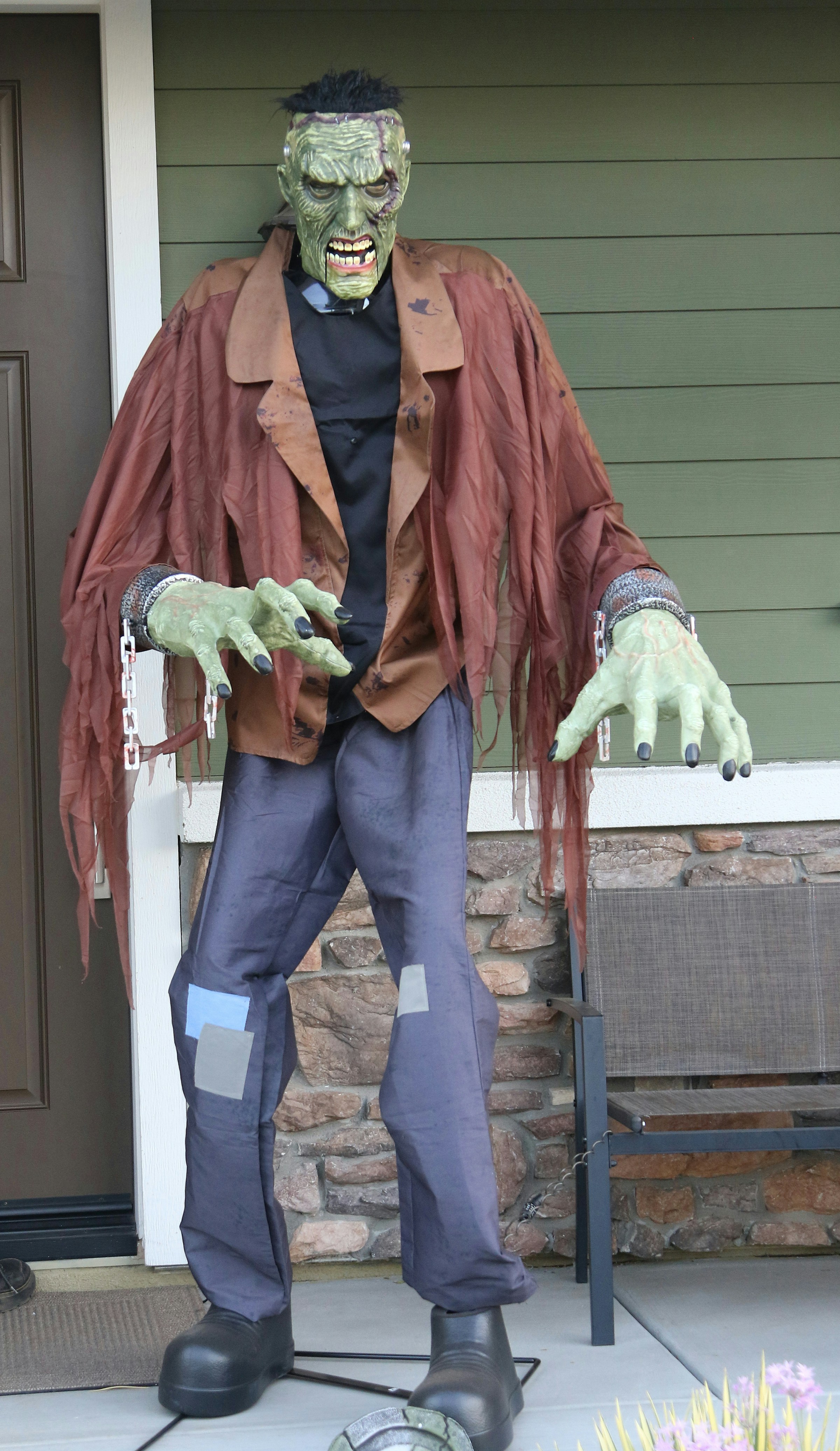 A green monster figure with chains on its wrists in front of a house