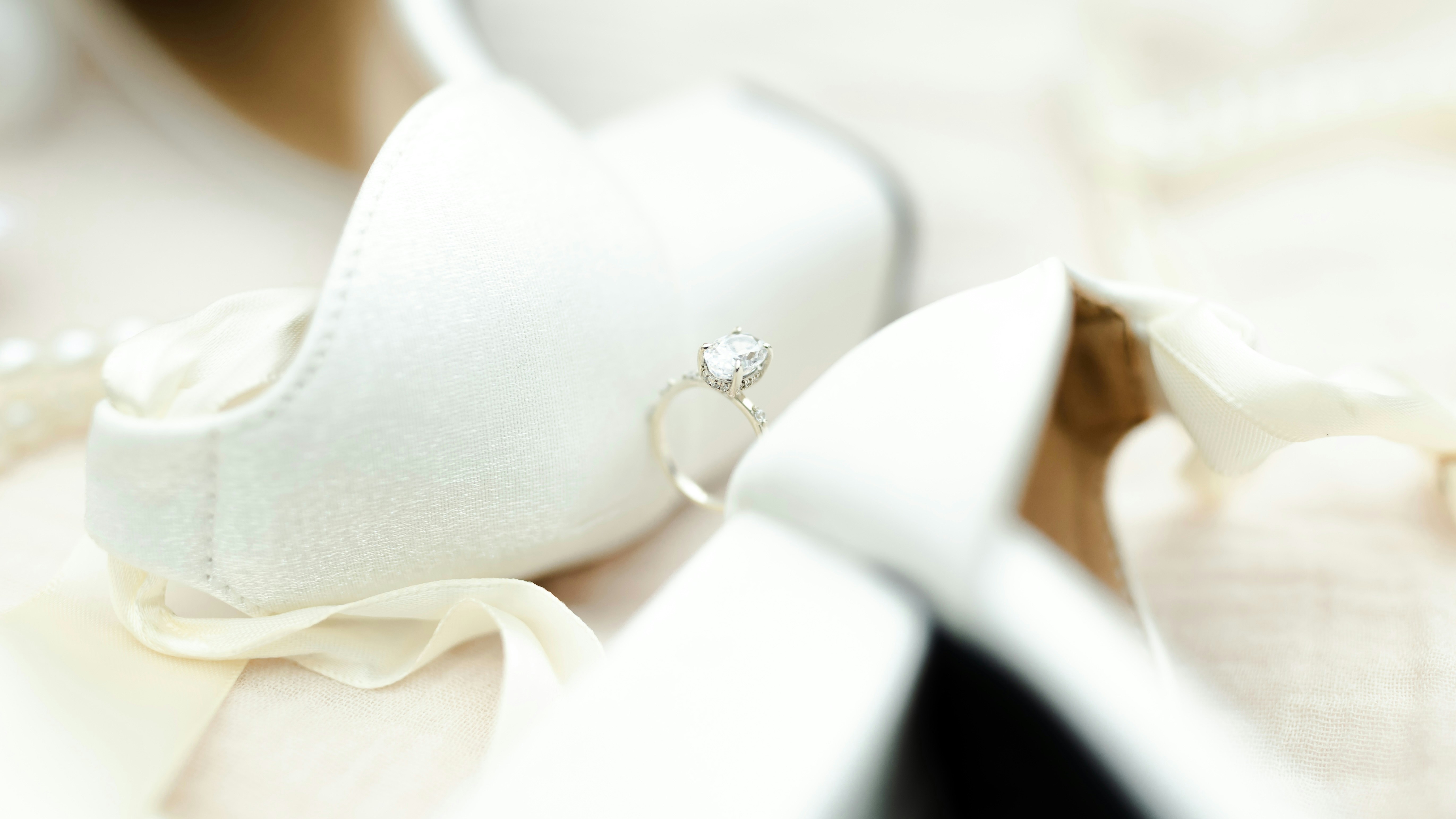 A diamond engagement ring resting between two white satin bridal shoes