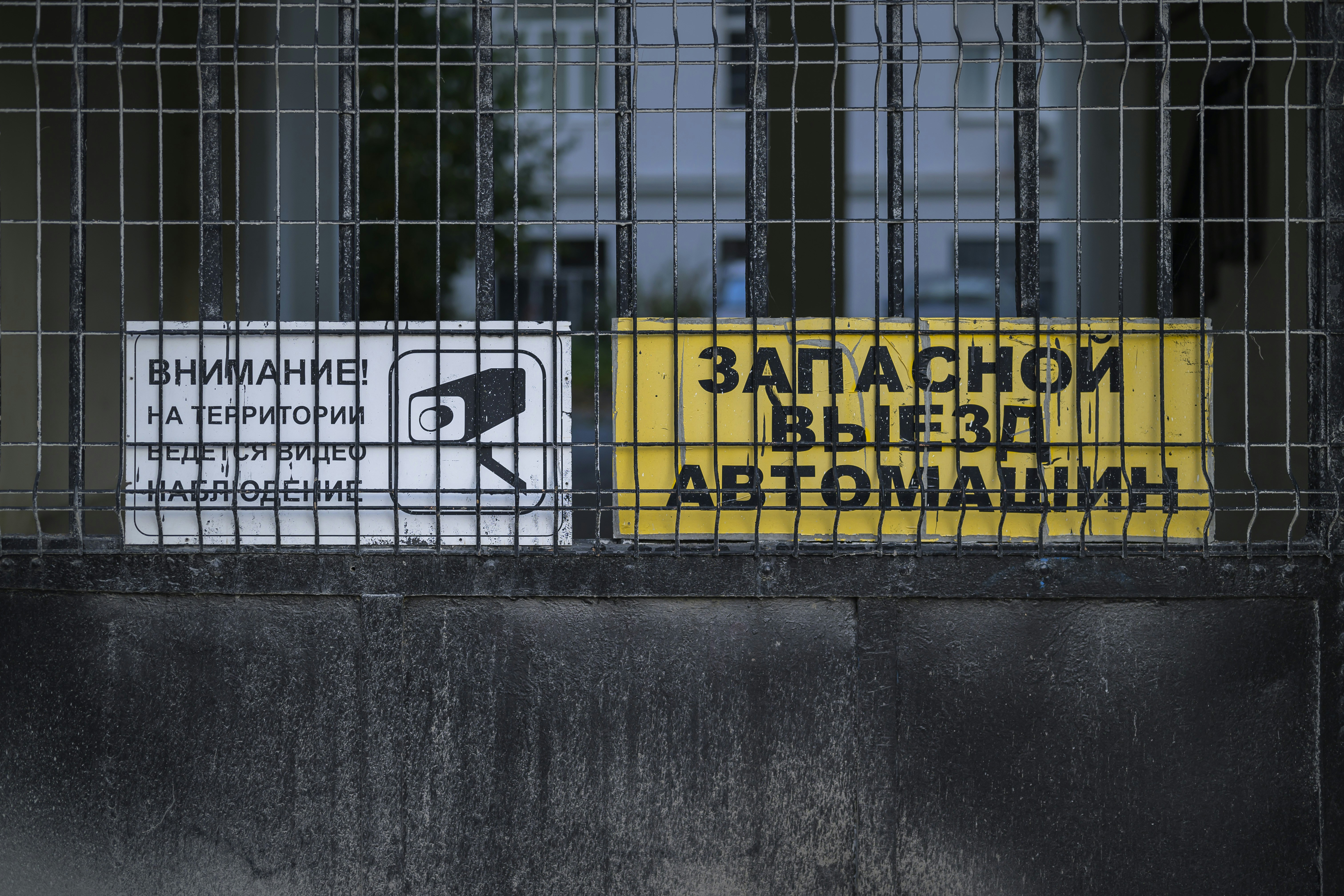 Carteles de advertencia blancos y amarillos en una verja metálica con un icono de cámara de seguridad