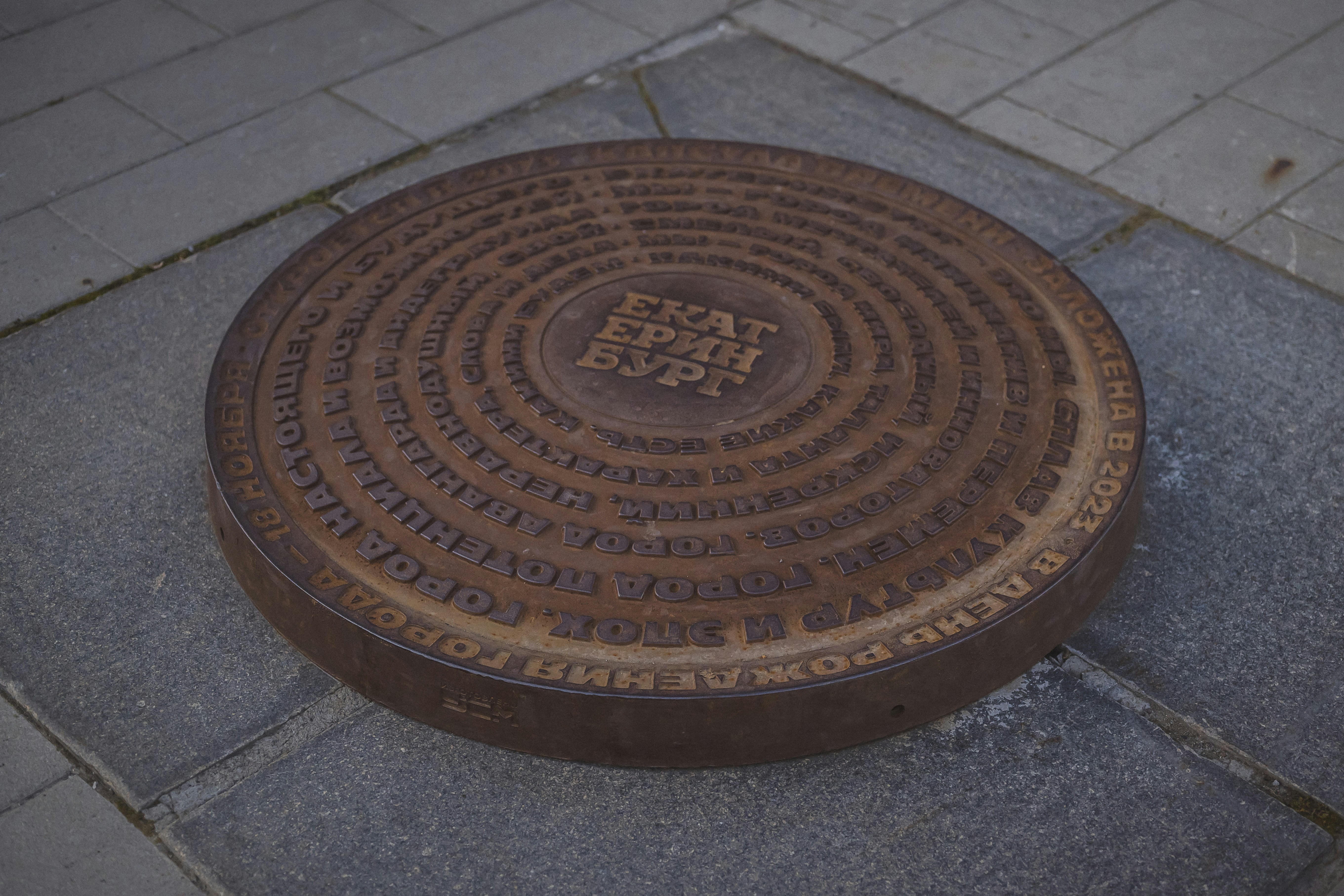 Una tapa circular de alcantarilla metálica con texto ruso en relieve sobre una acera de piedra