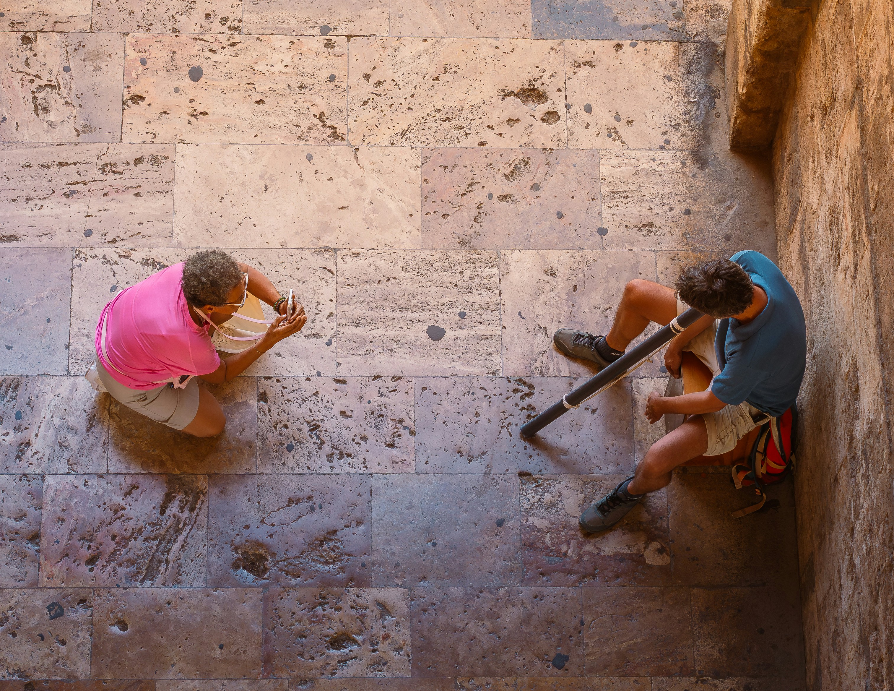 A person in a pink shirt photographing a person playing a didgeridoo on stone tiles