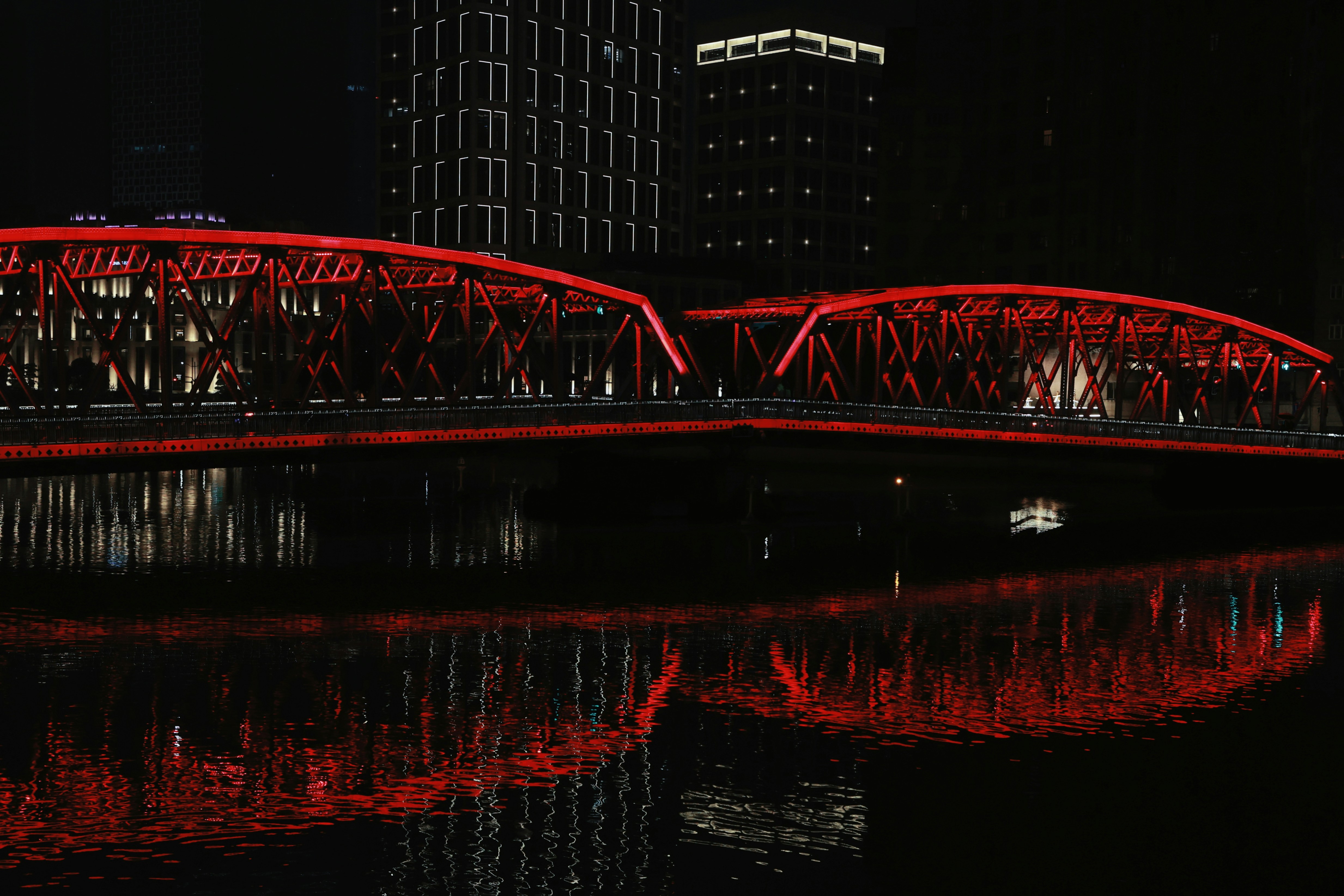 A red illuminated truss bridge reflecting in dark water at night