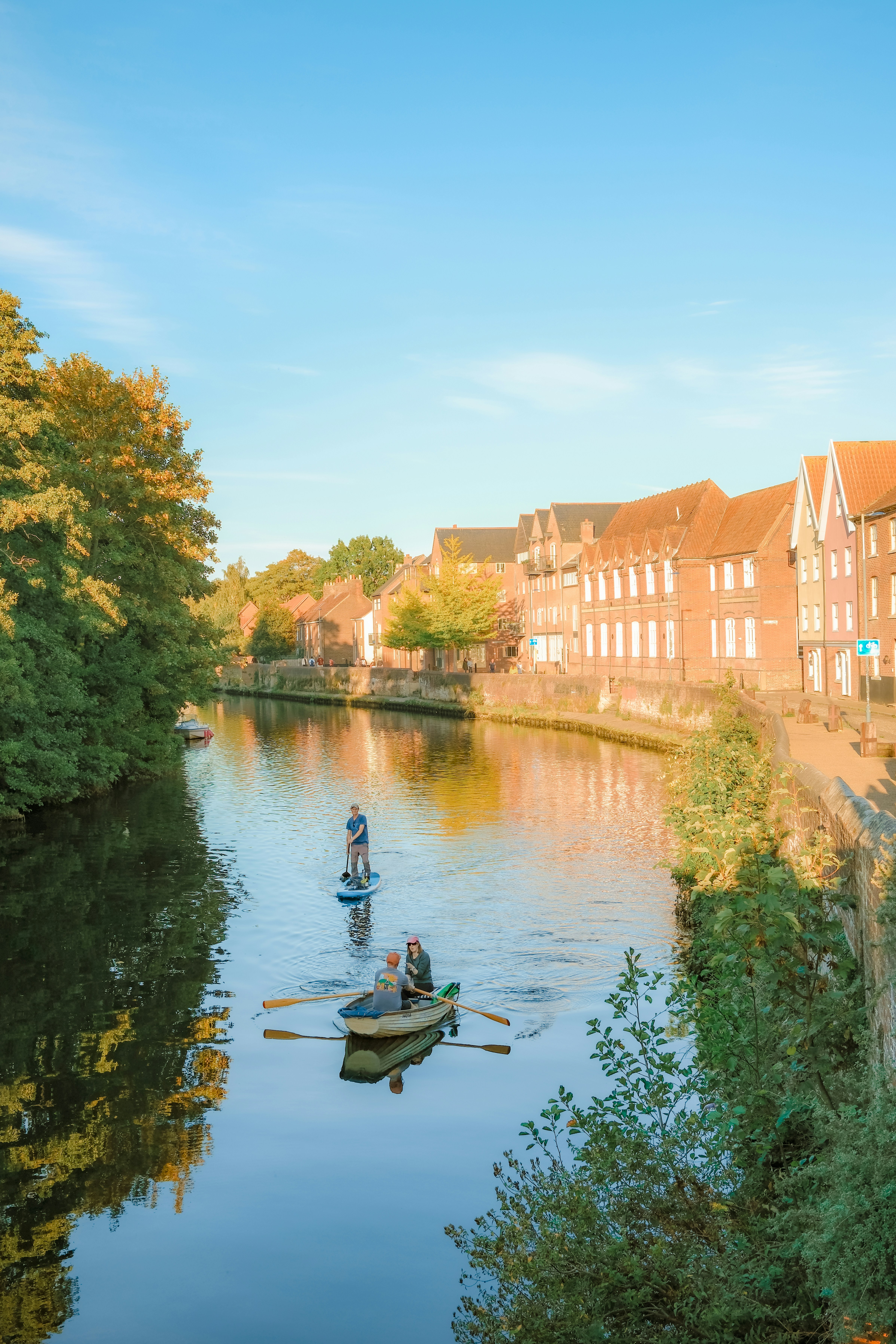 People rowing a boat and paddleboarding on a river beside brick buildings