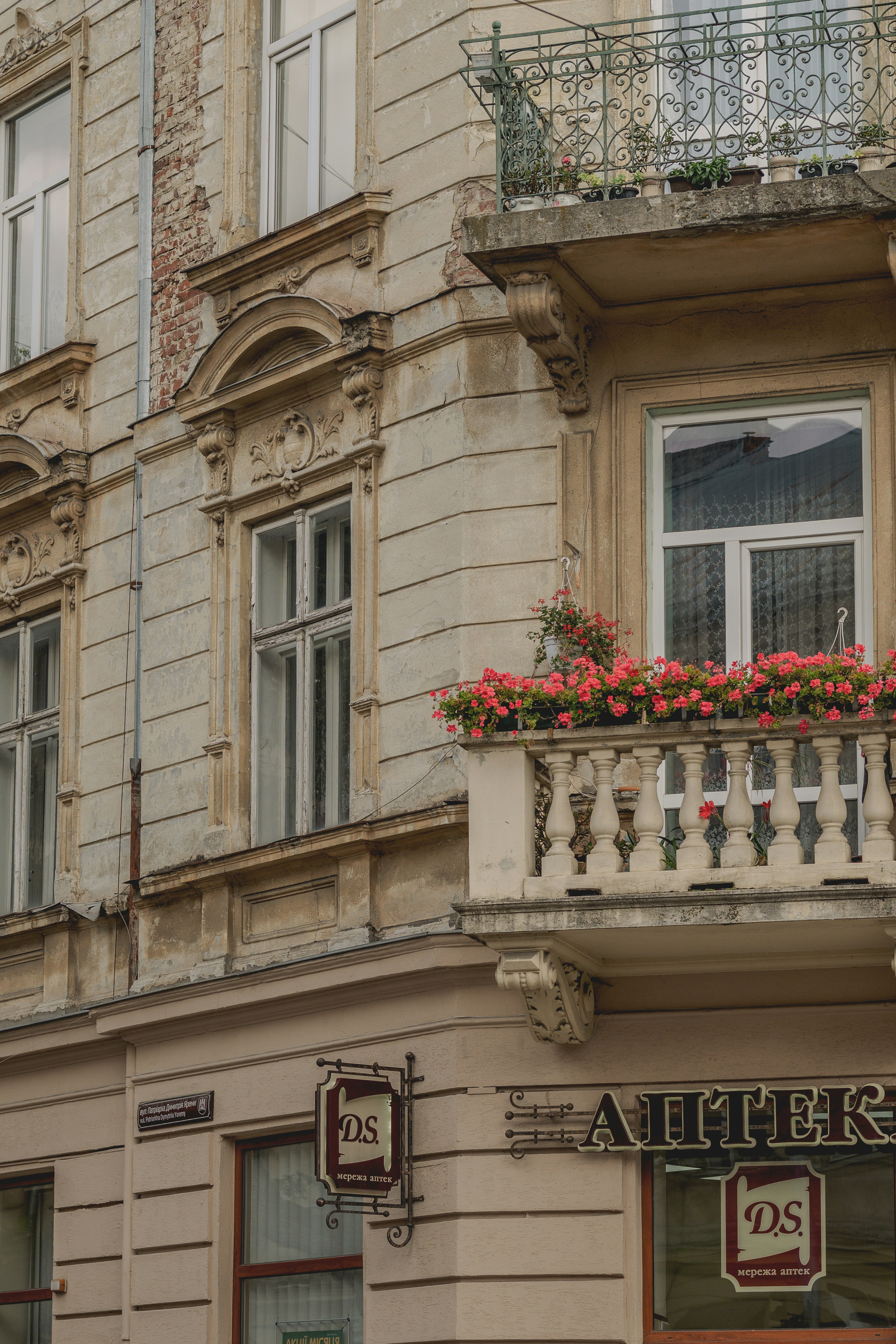A stone building facade with a balcony featuring pink flowers and a pharmacy sign