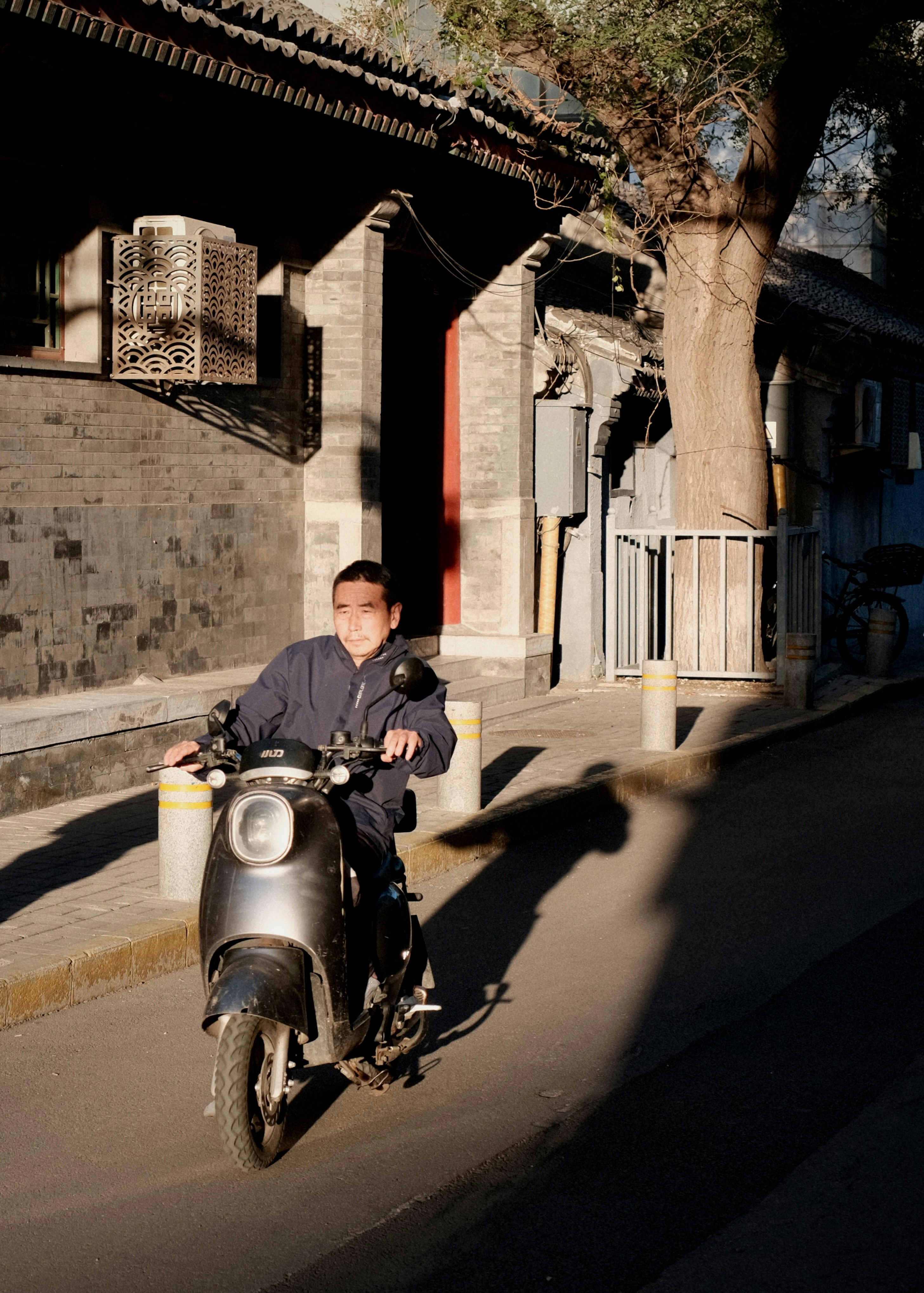 A man riding a grey electric scooter past a traditional brick building in Beijing