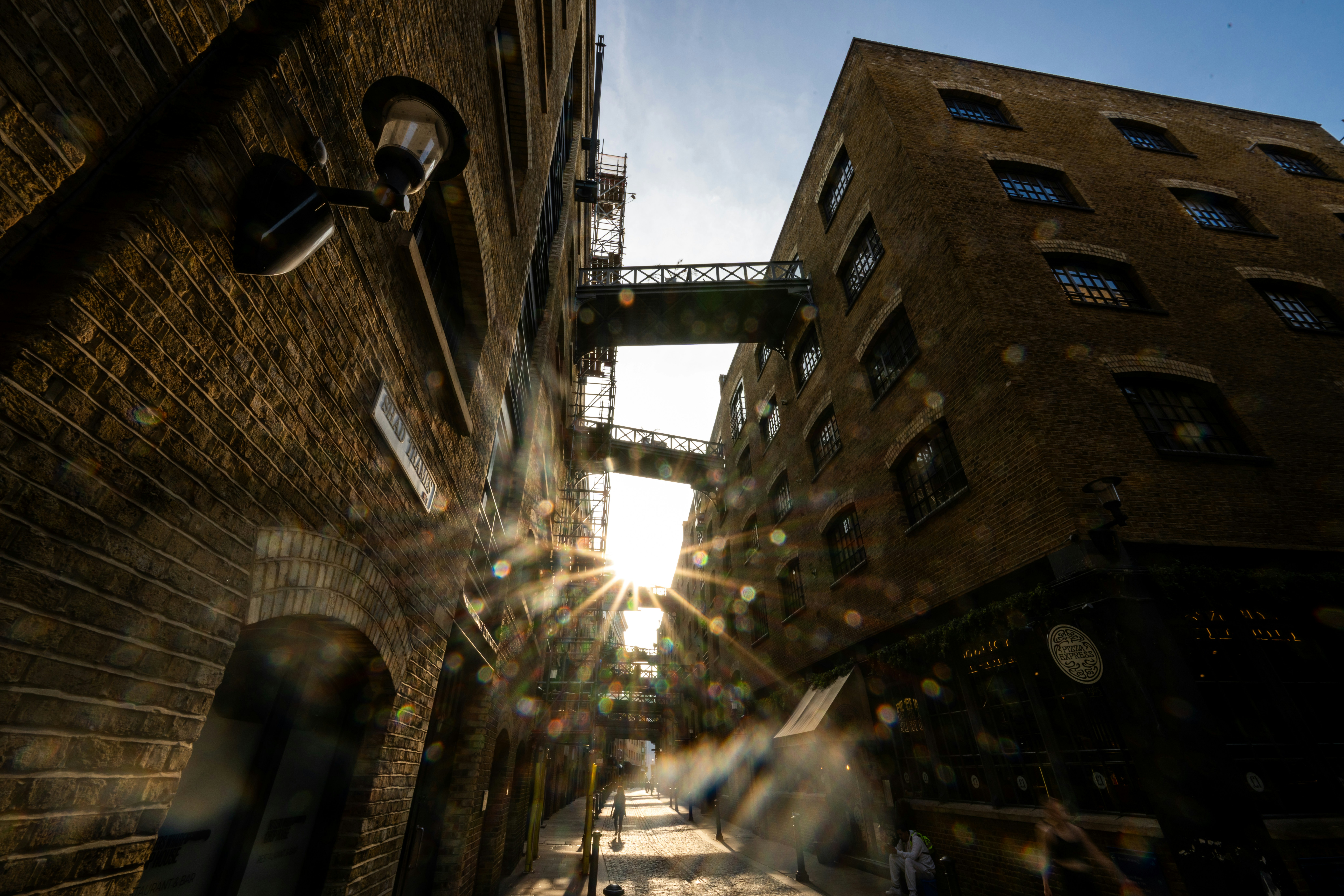 Low sun shining between tall brick warehouse buildings connected by metal walkways in London