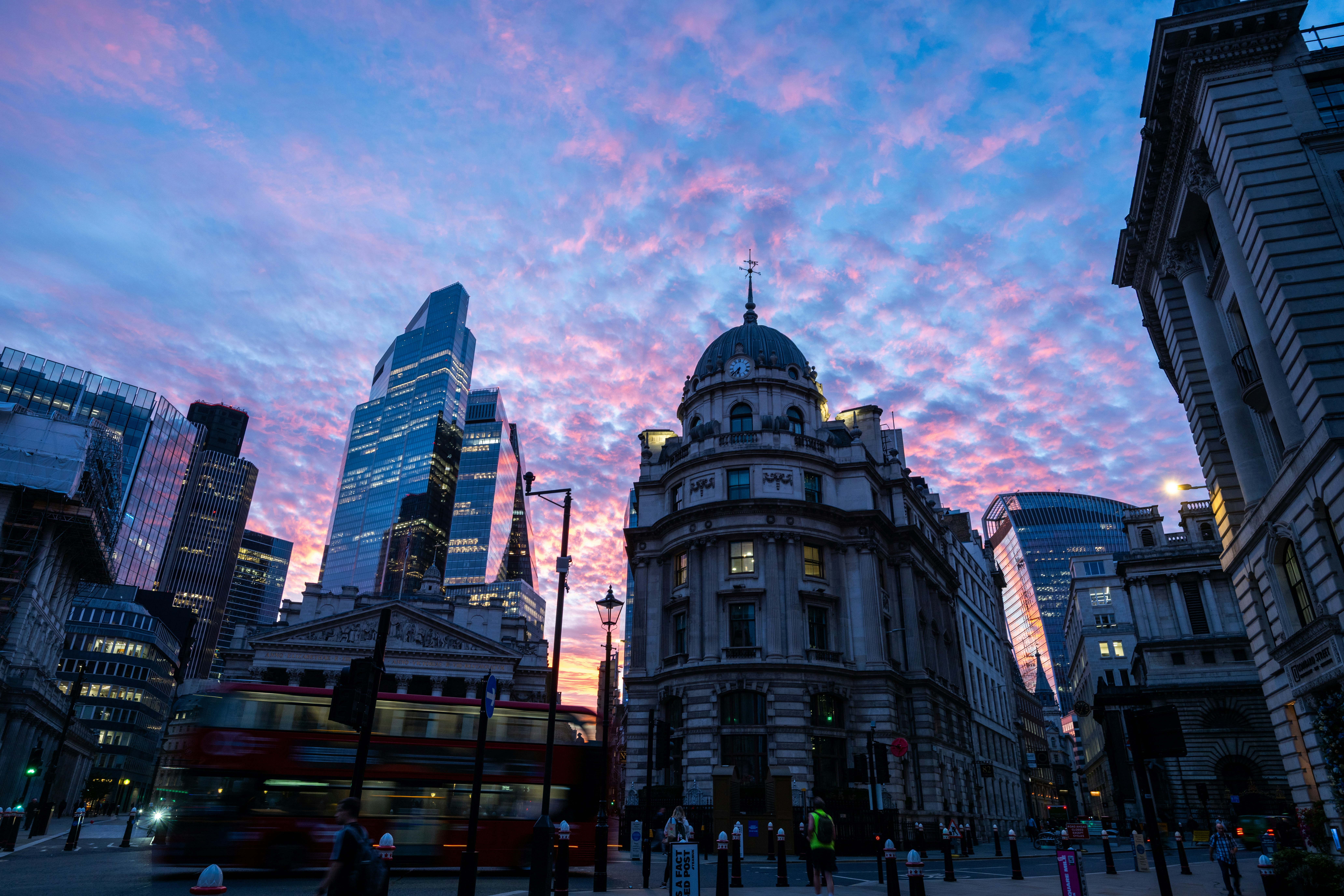 A red double-decker bus moving past historic buildings under a vibrant pink and blue sky