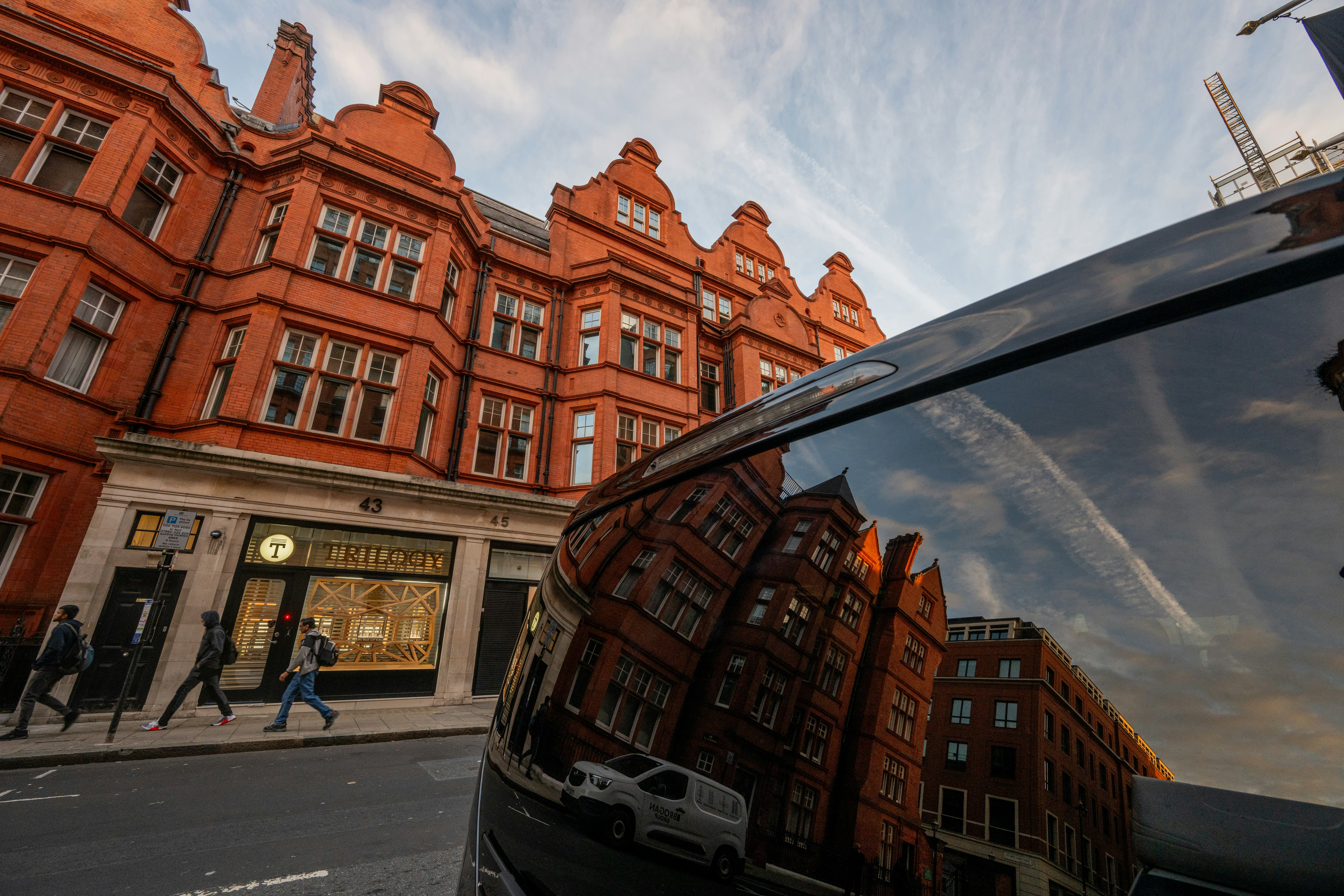 People walking past red brick buildings with reflections on a car surface