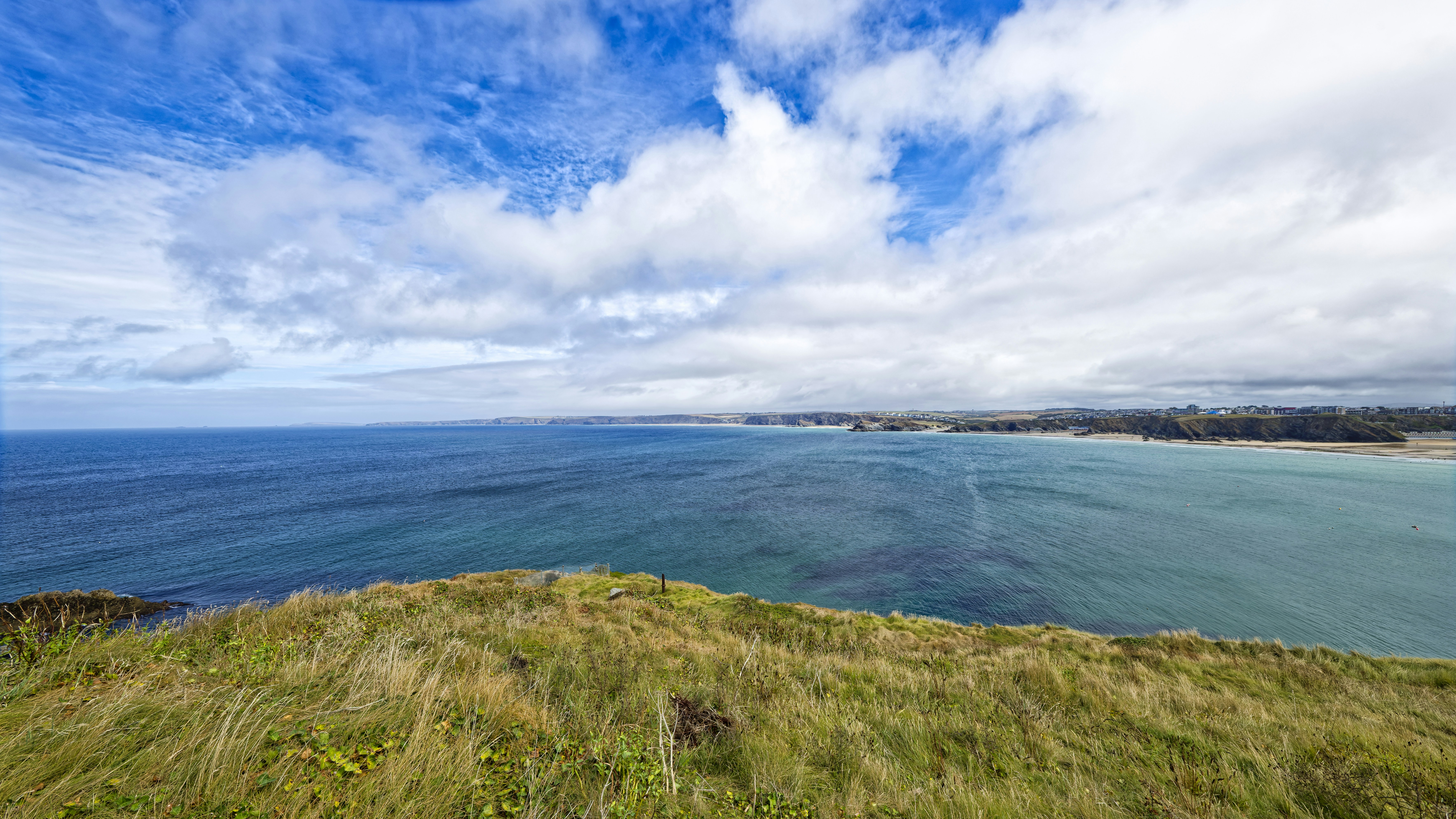 A grassy cliff overlooking a blue bay with a sandy beach under clouds