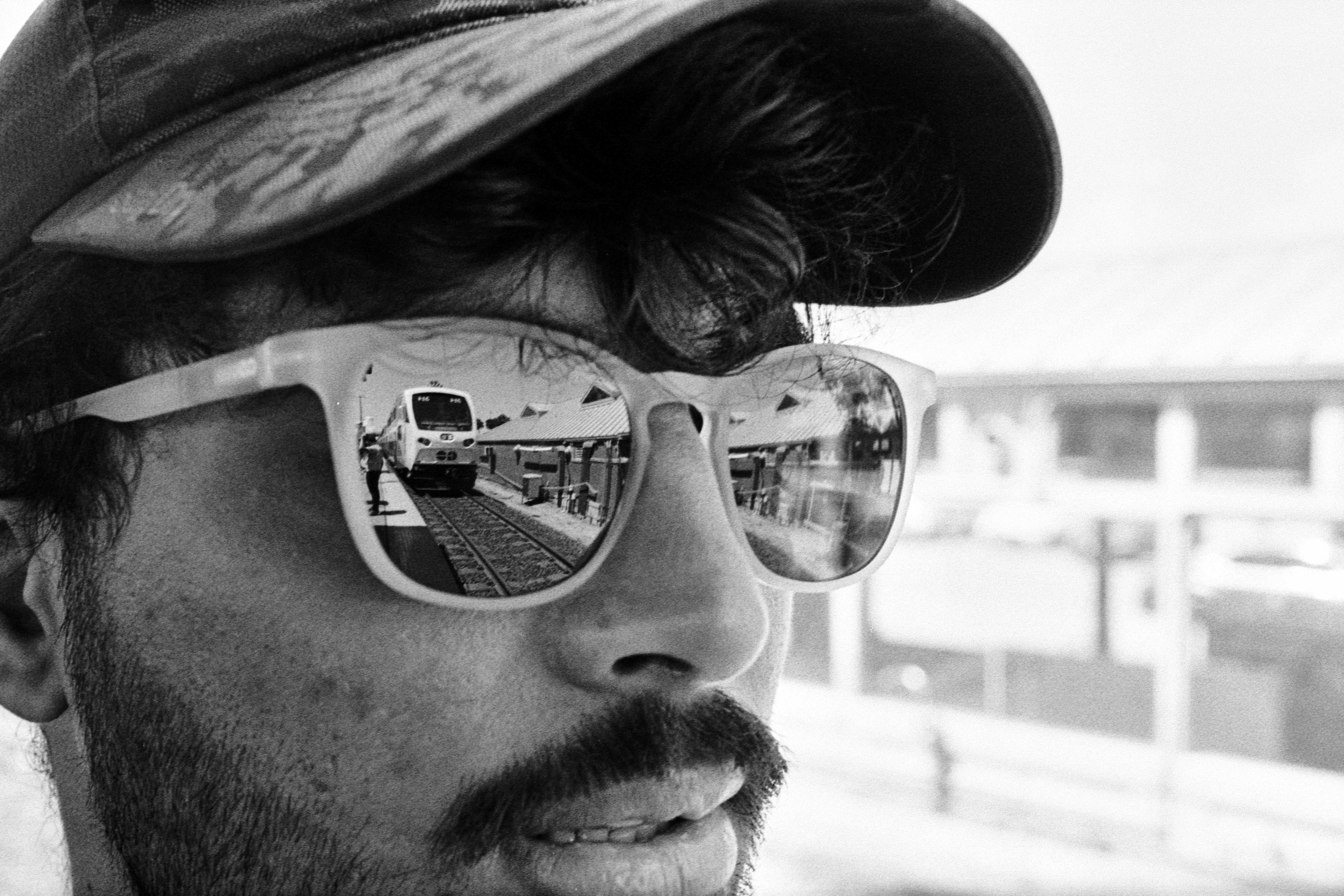 A person wearing a baseball cap and sunglasses reflecting a train at a station