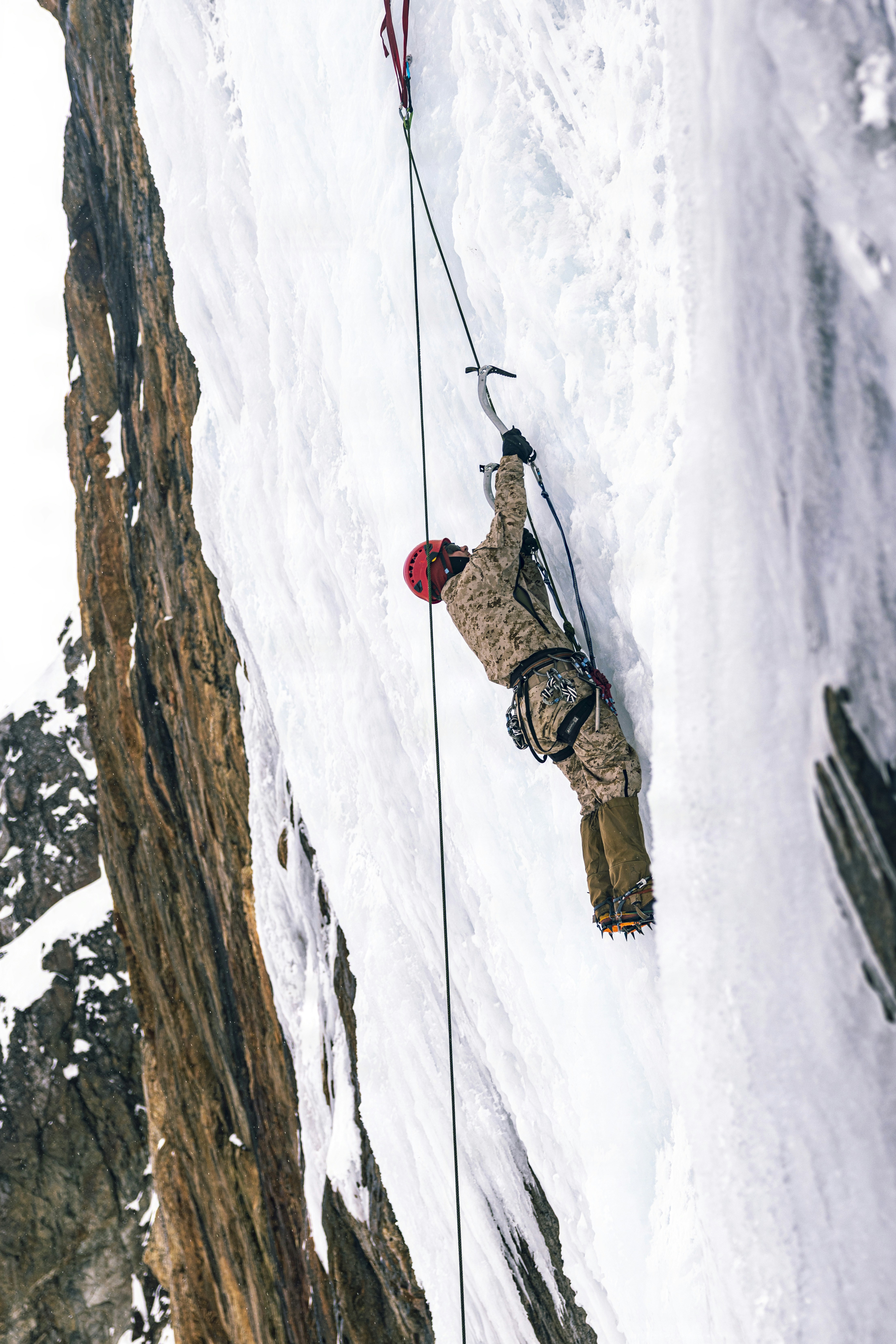 A person in camouflage gear ice climbing a frozen waterfall on a cliff