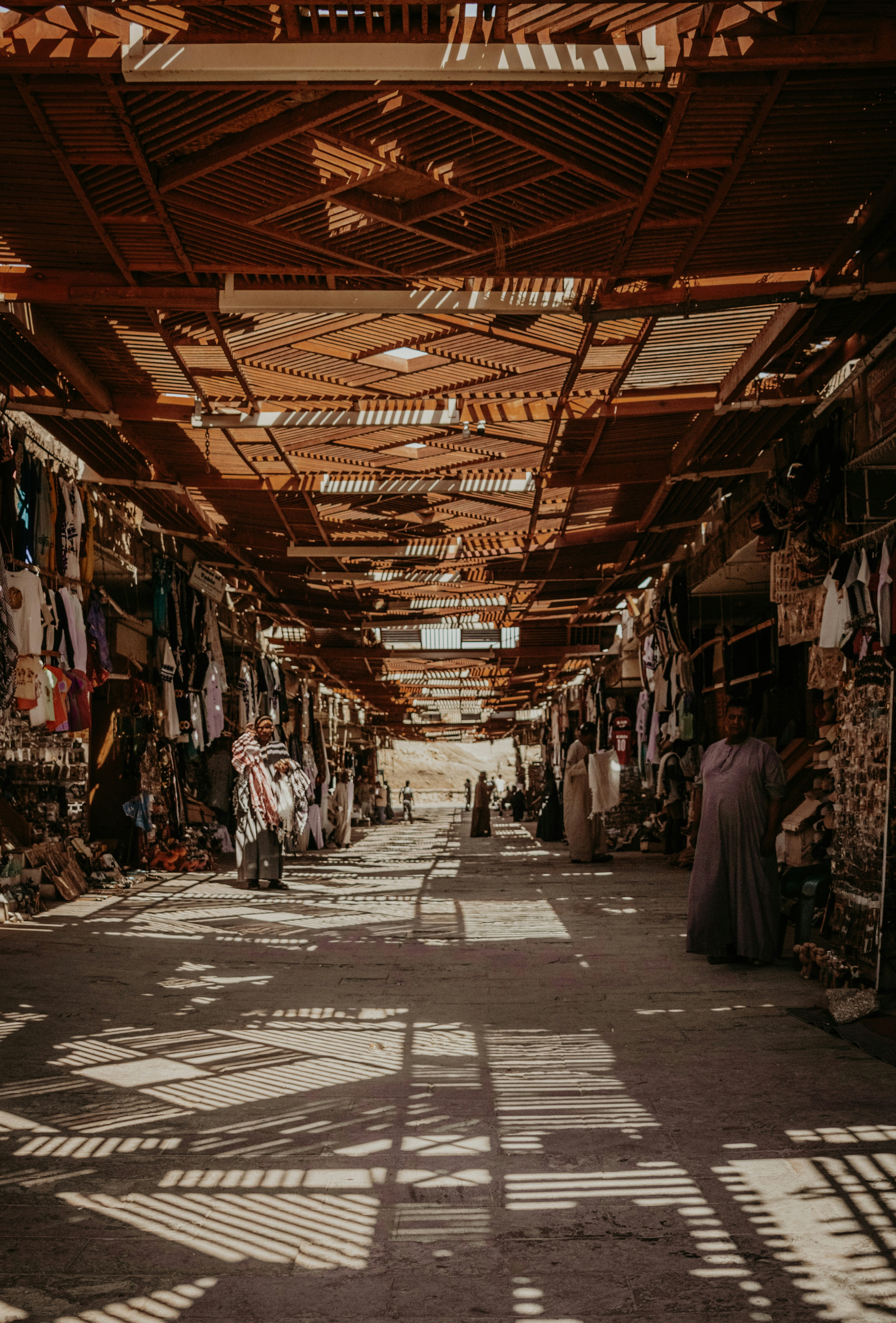 People in a traditional market with a slatted wooden roof casting long shadows
