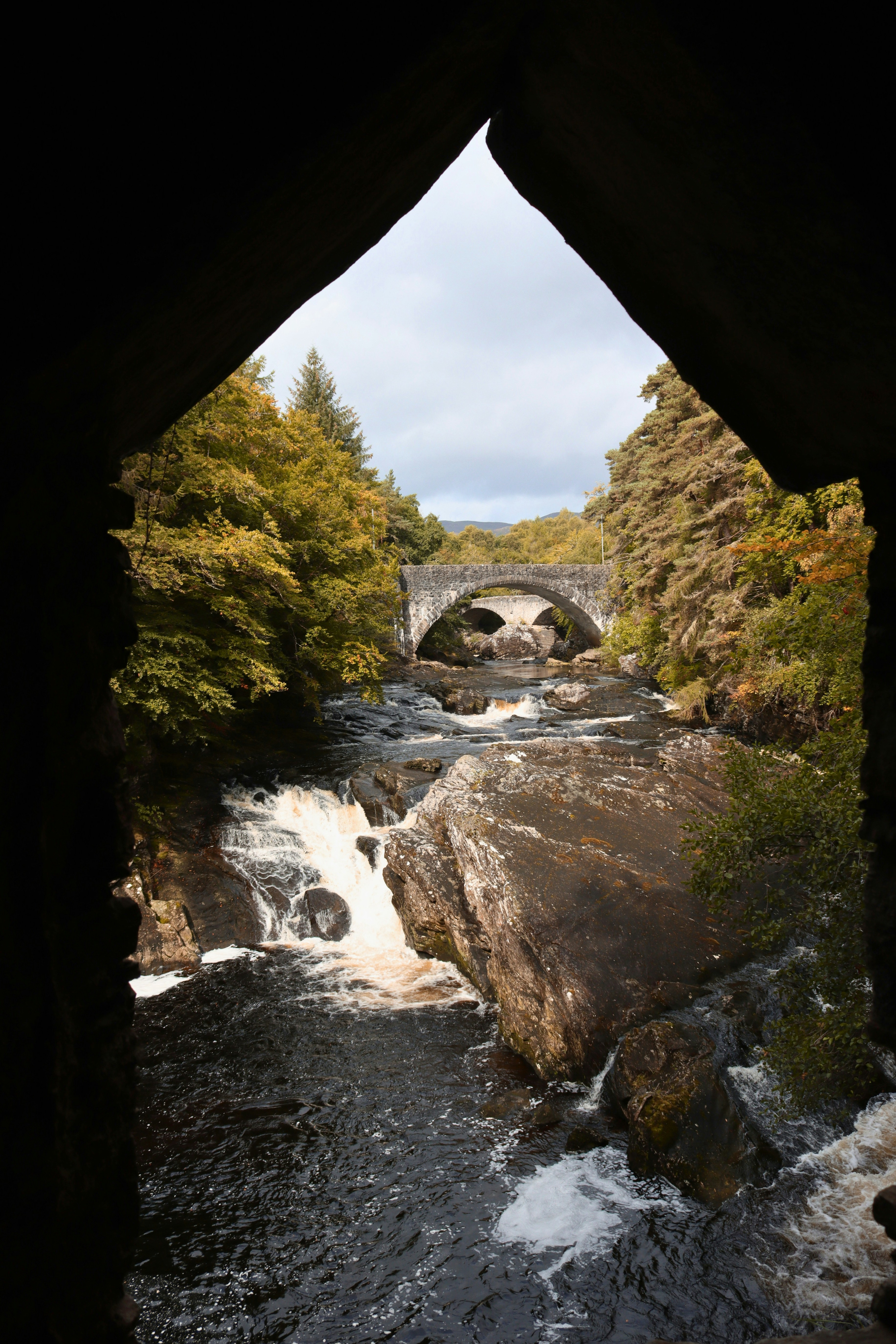 A stone bridge over a rocky river with waterfalls framed by a dark archway