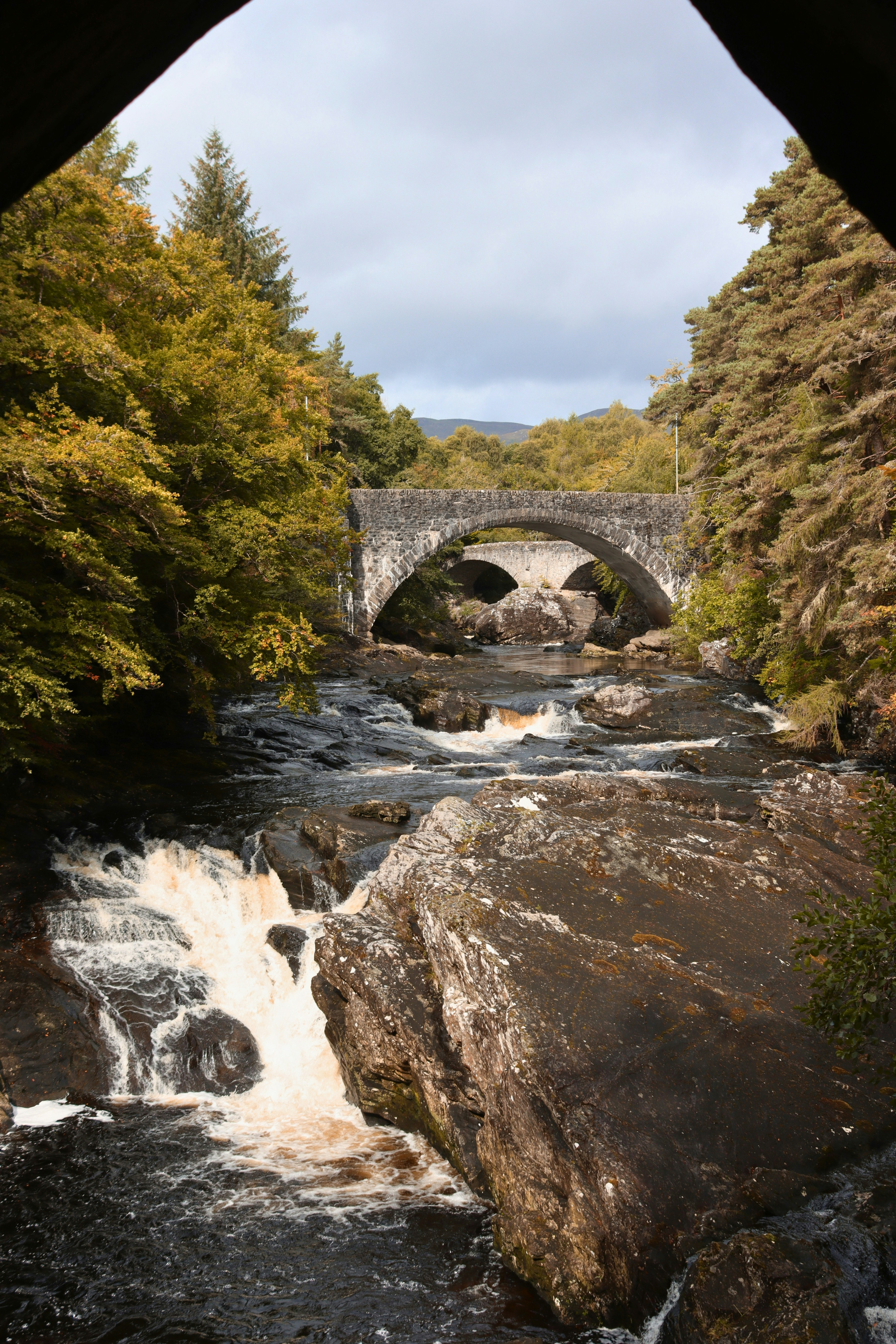 A stone arch bridge over a rocky river with small waterfalls and trees