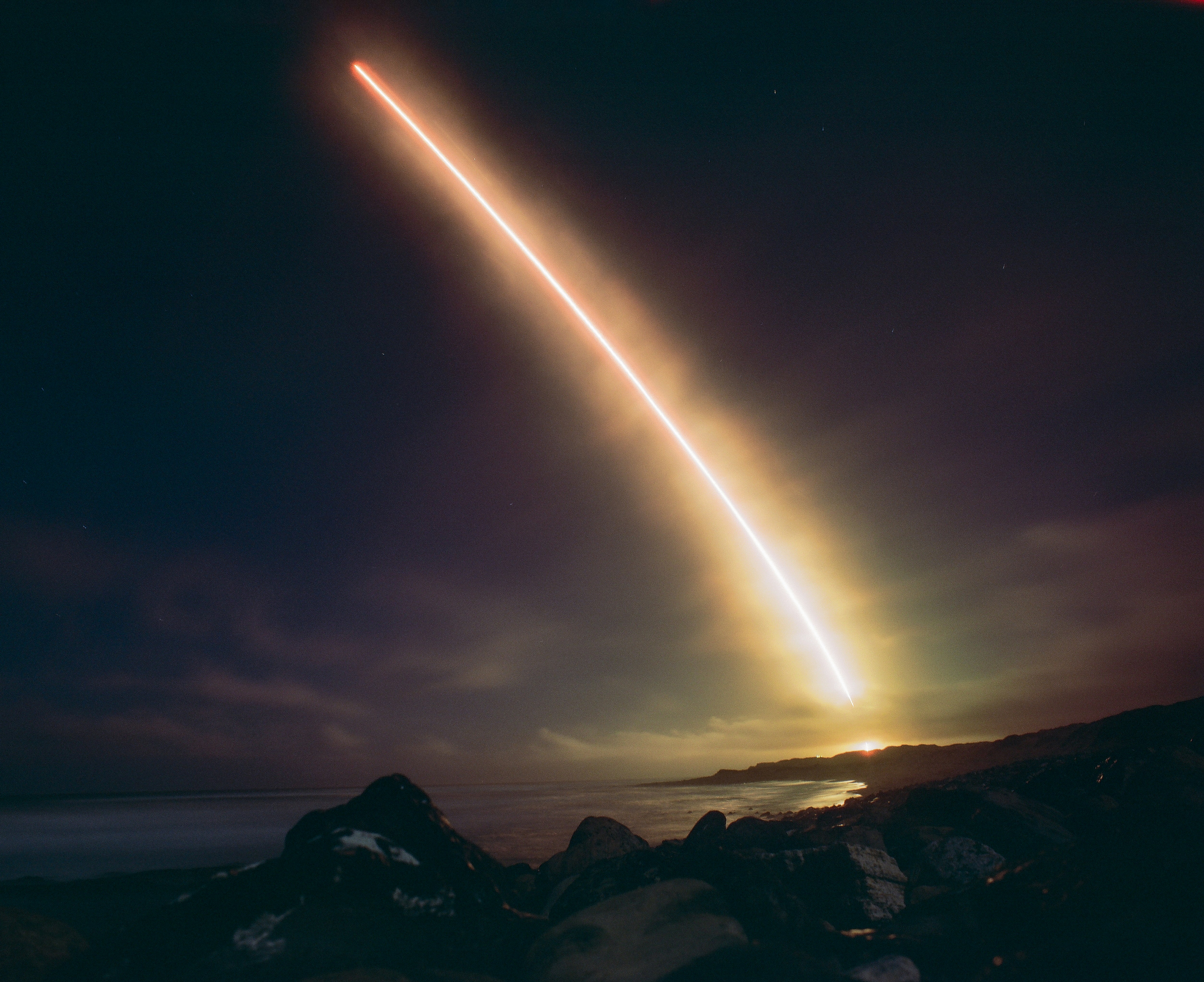 A bright light streak across a dark night sky above a rocky coastline