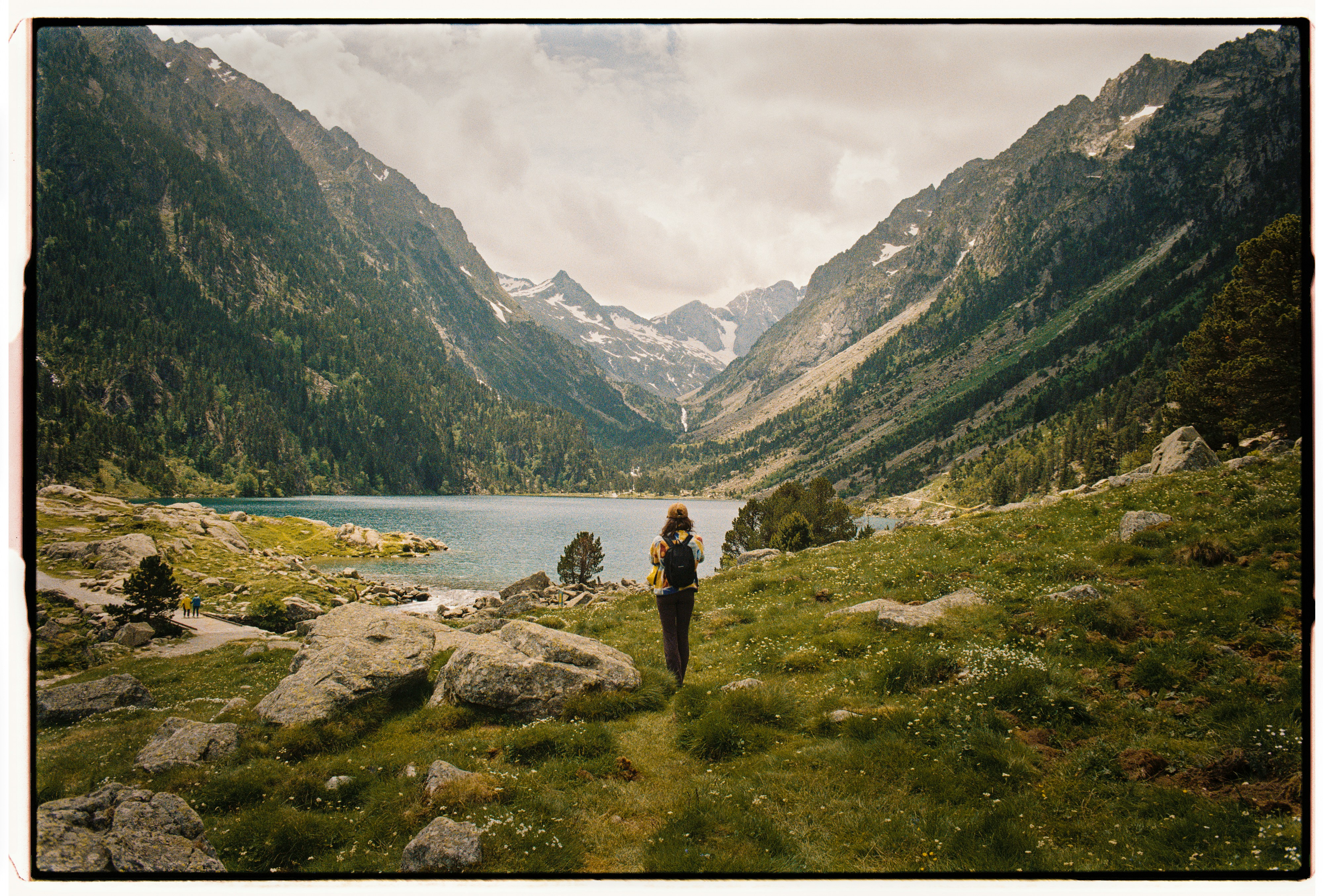 A person with a backpack walking toward a mountain lake in a valley