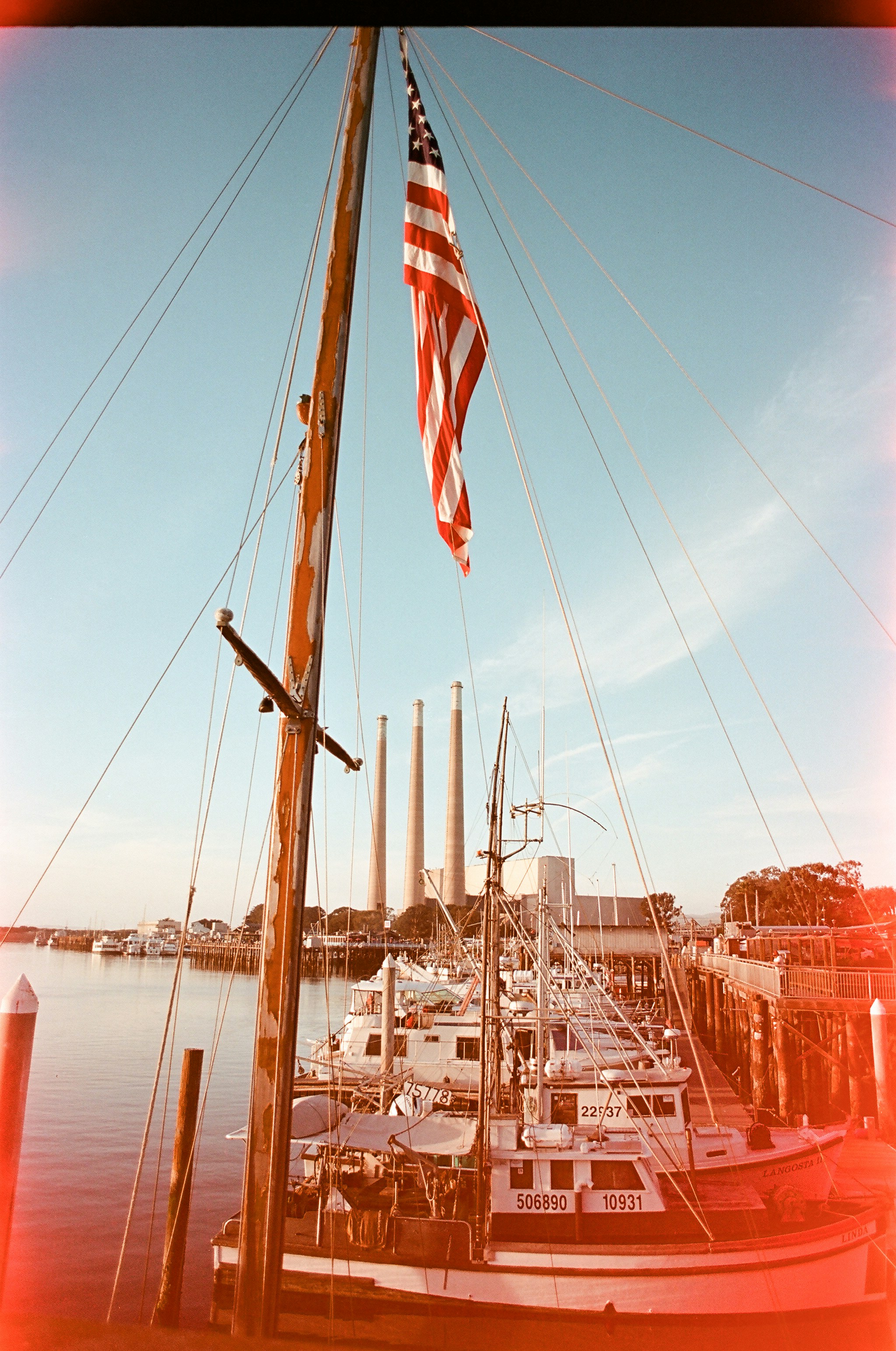An American flag flying above fishing boats docked at a pier near power plant smokestacks