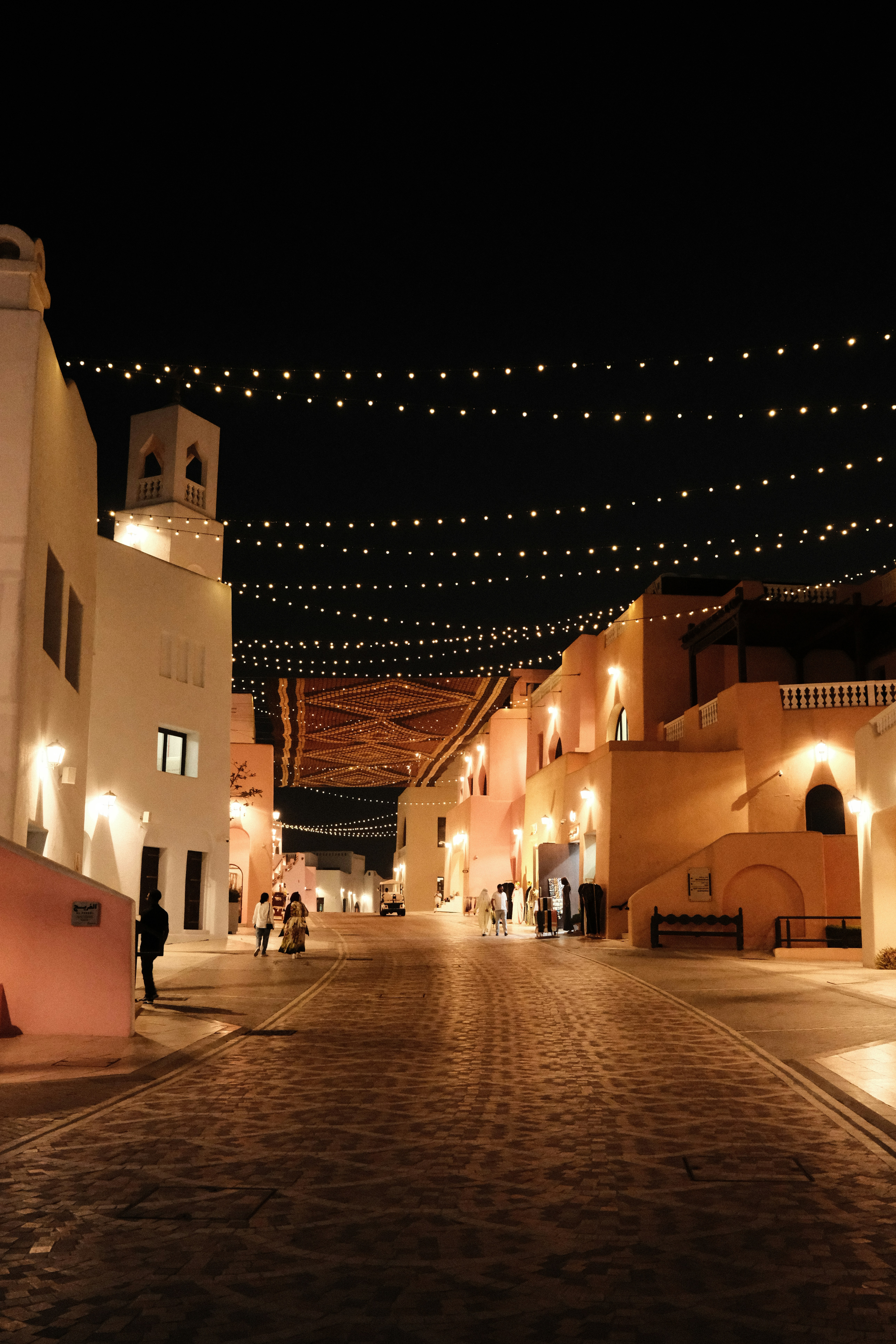 People walking on a cobblestone street under strings of glowing lights at night