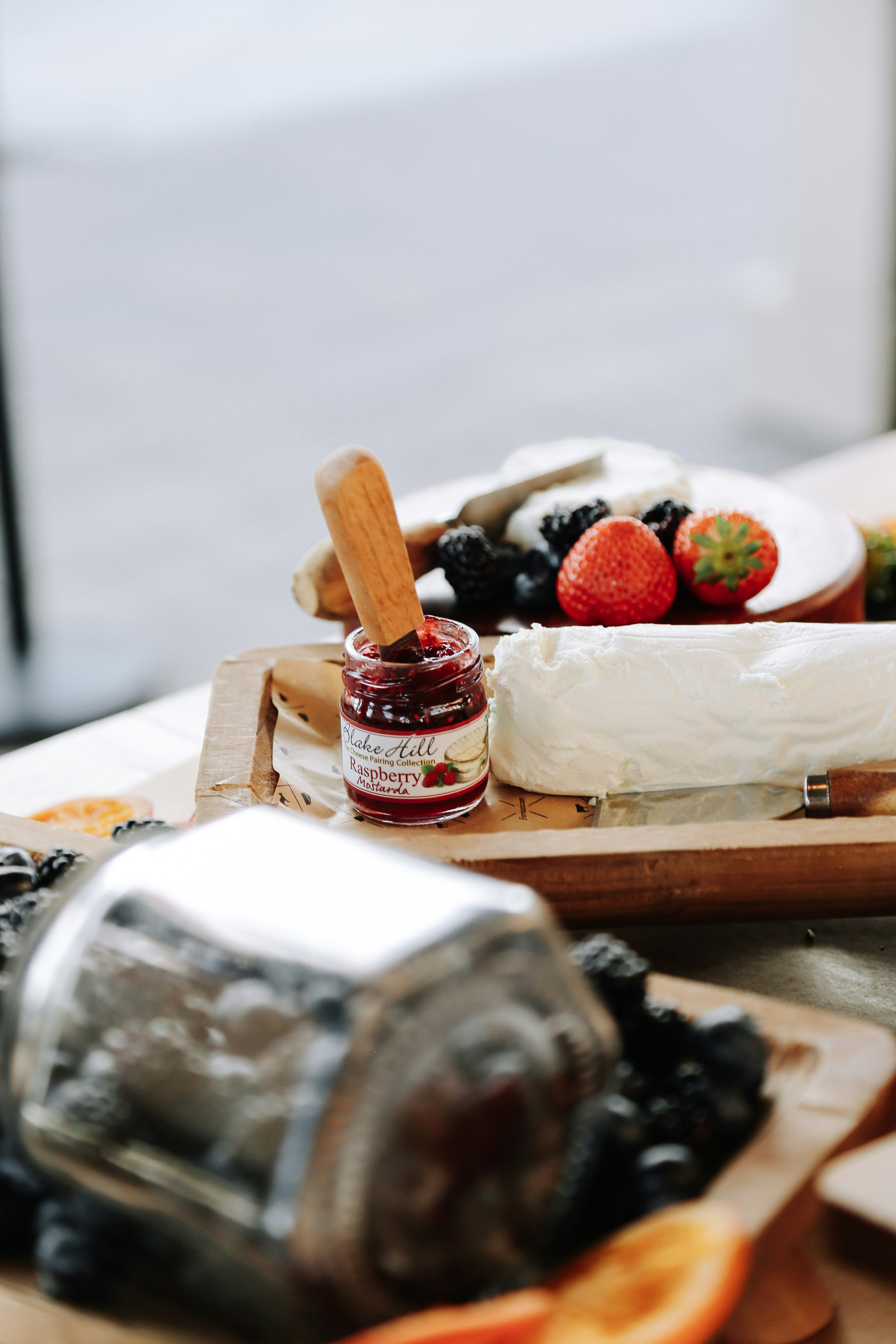 A jar of raspberry jam with a wooden spreader beside goat cheese and berries