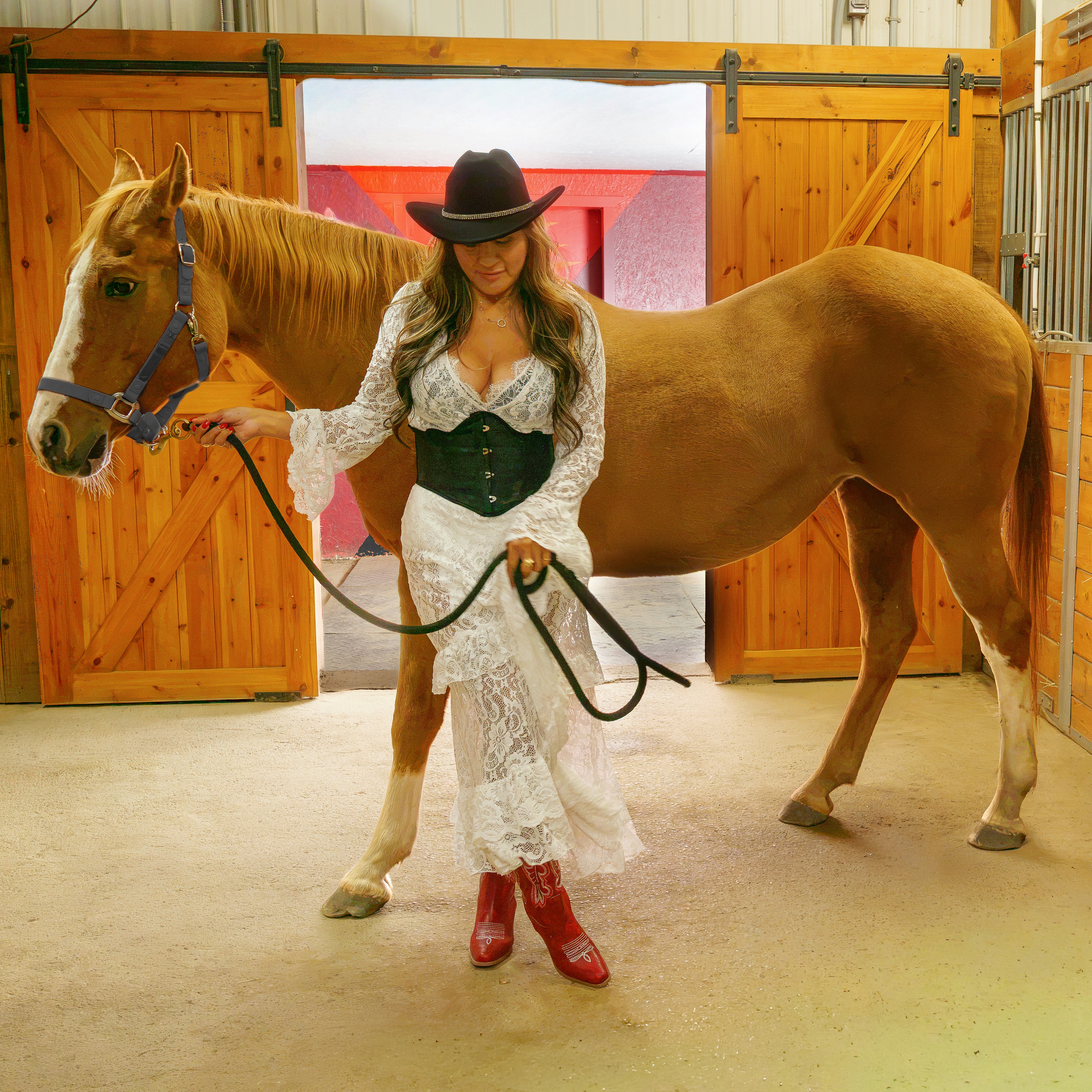 A woman in a white lace dress holding a brown horse in a stable