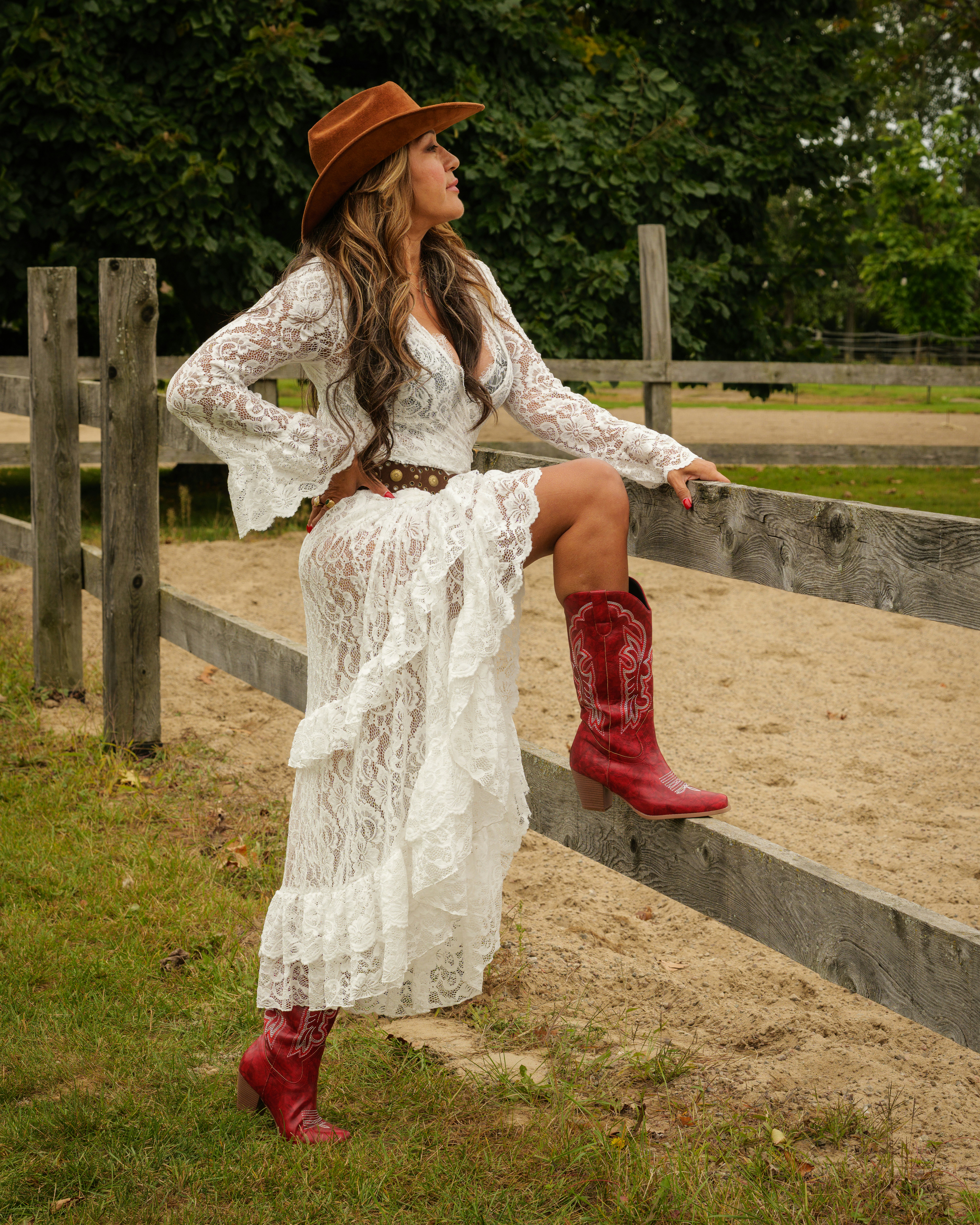 Uma mulher com um vestido branco de renda e botas de cowboy vermelhas ao lado de uma cerca de madeira