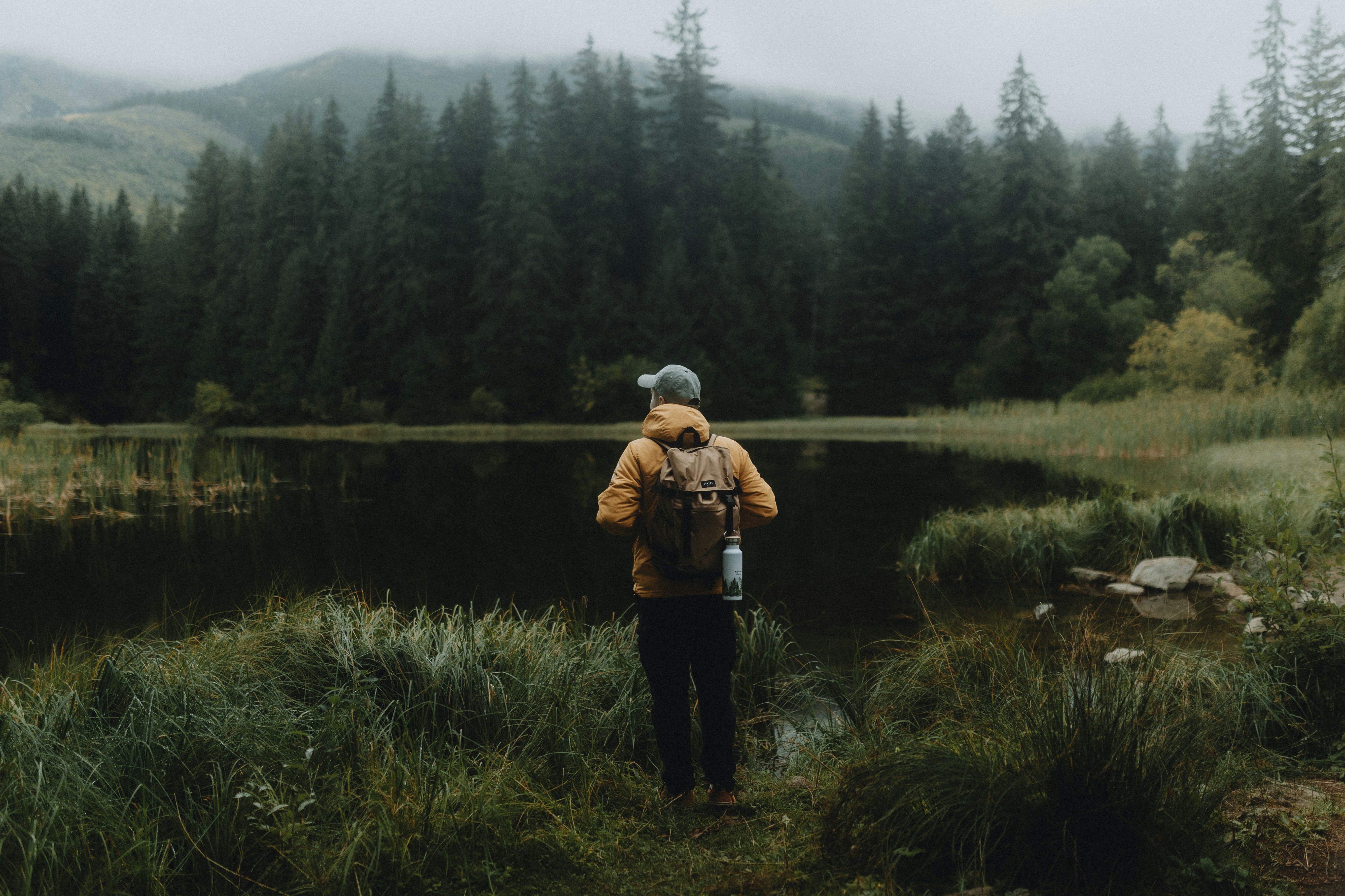A person with a backpack standing by a dark lake near a pine forest