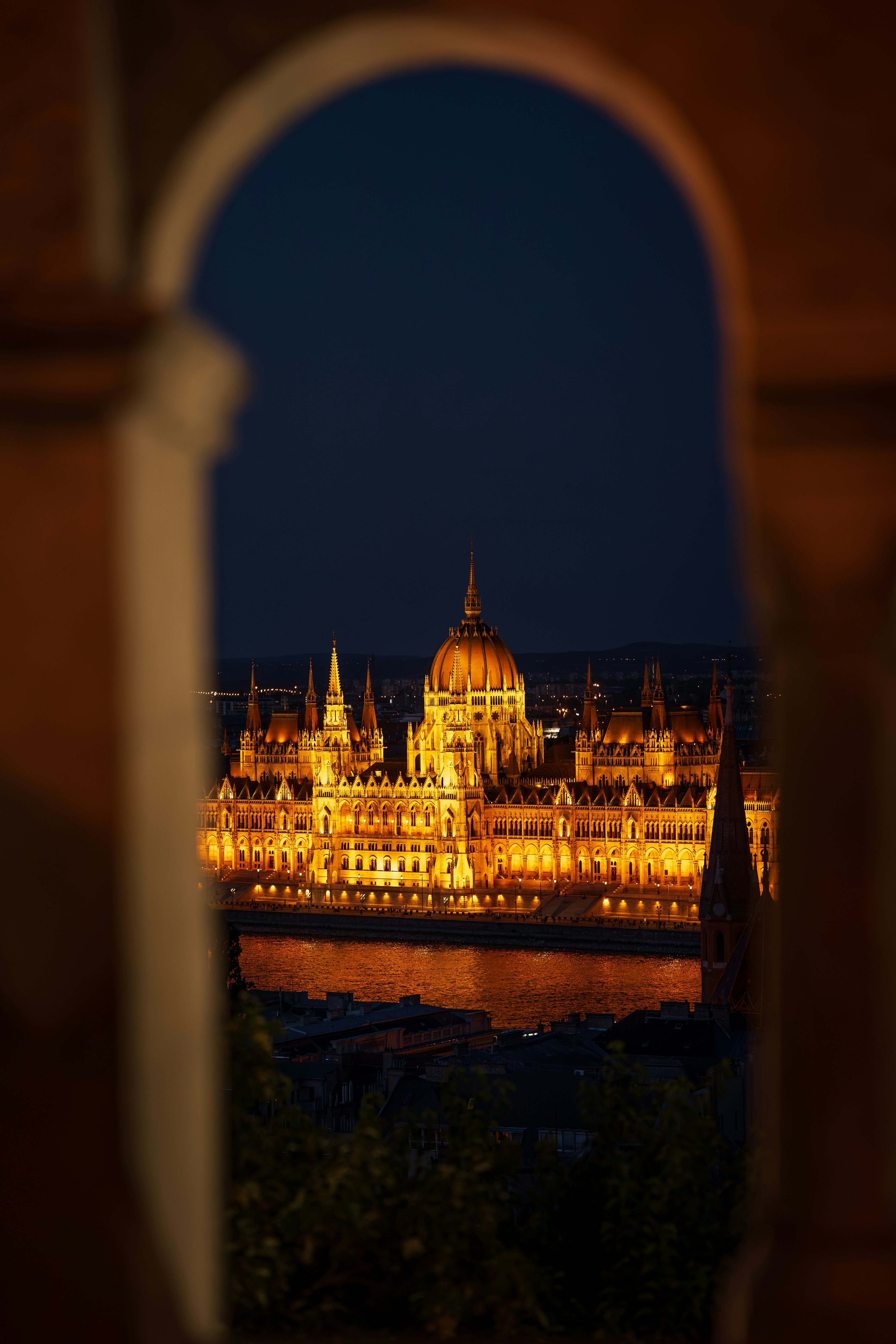 The Hungarian Parliament Building illuminated at night, viewed through a stone archway in Budapest