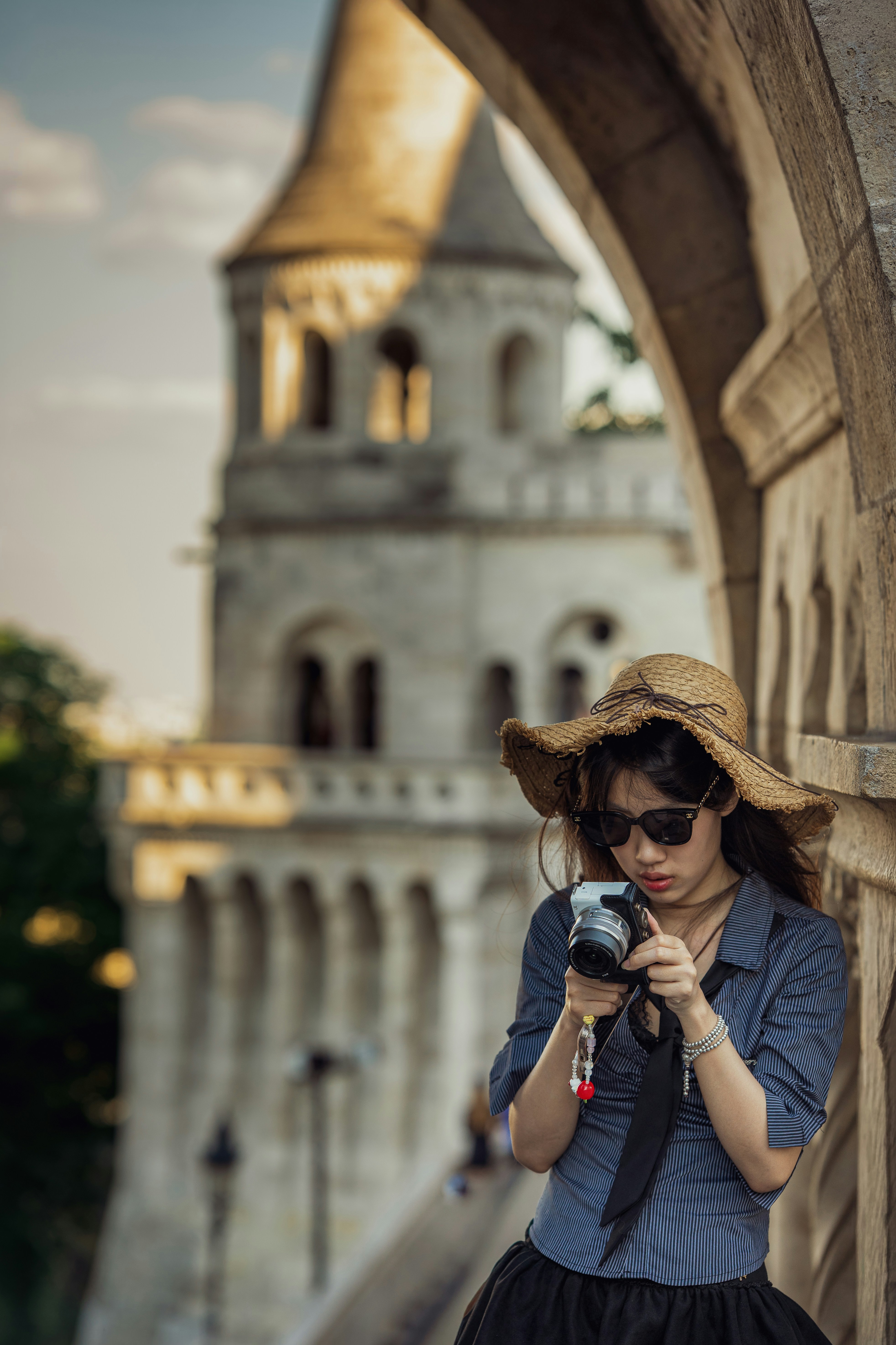 A woman in a straw hat and sunglasses holding a camera before a stone tower