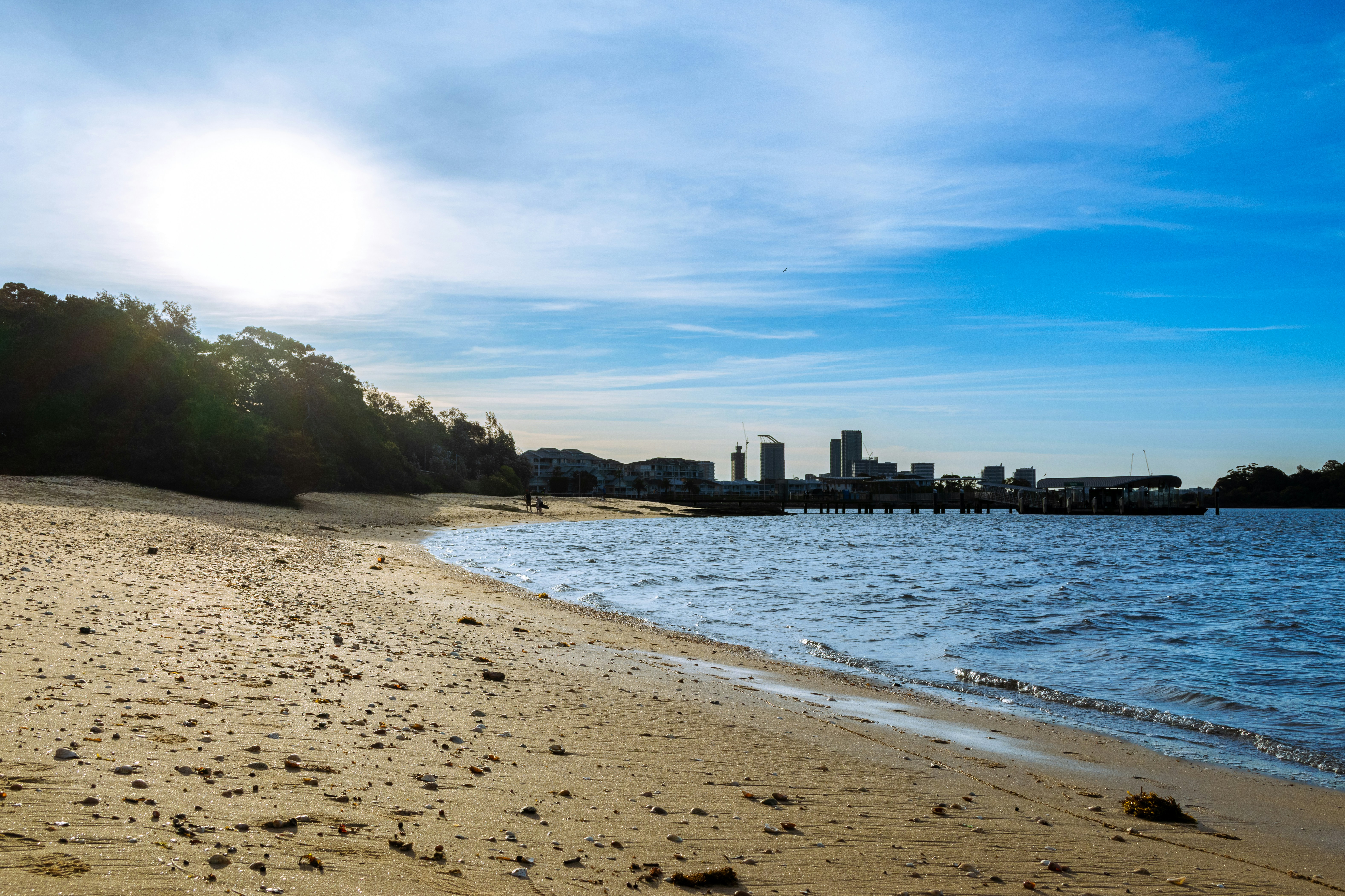 A sandy beach with a pier and distant city skyline under a bright sky