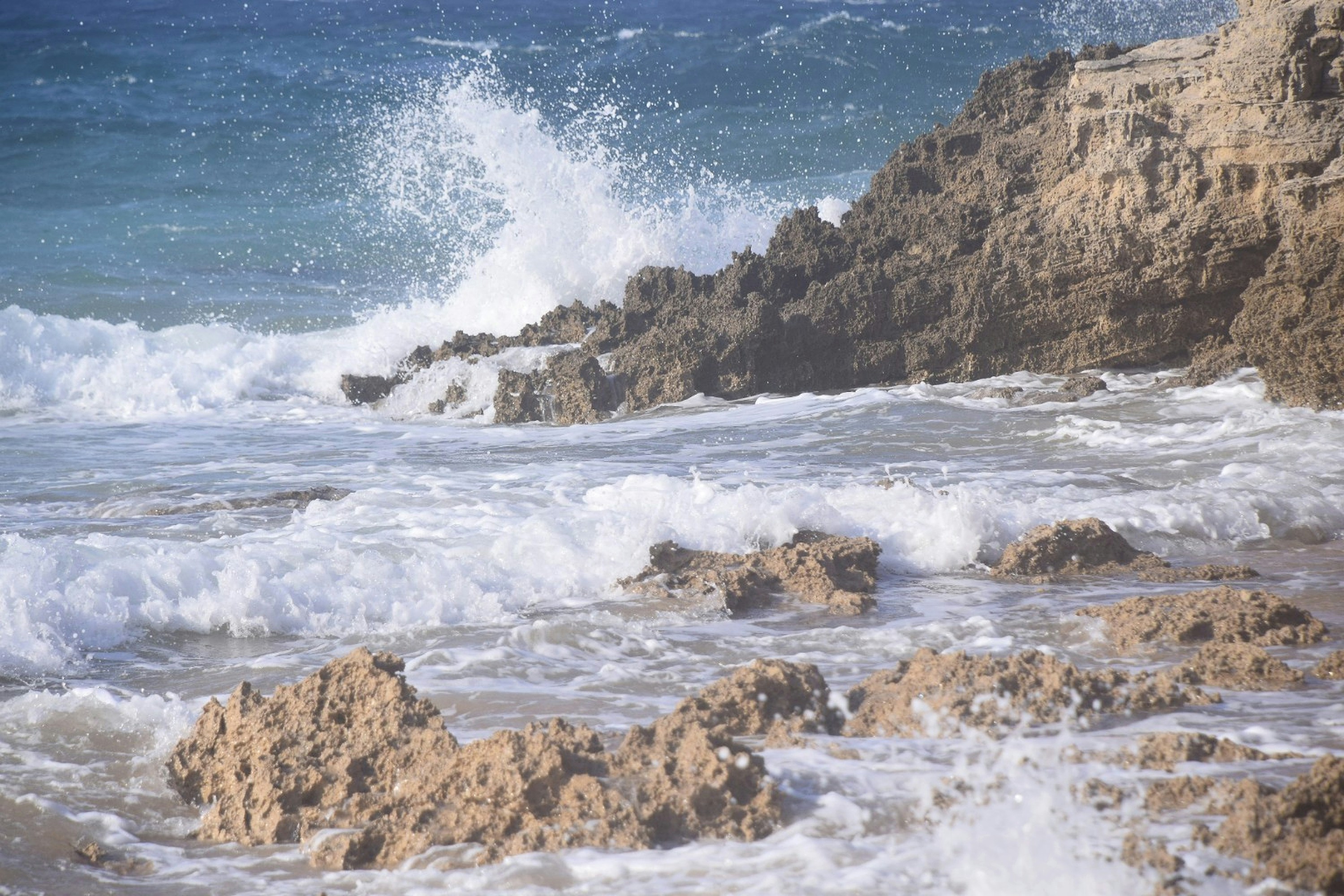 Ocean waves crashing against jagged brown rocks on a sandy shore