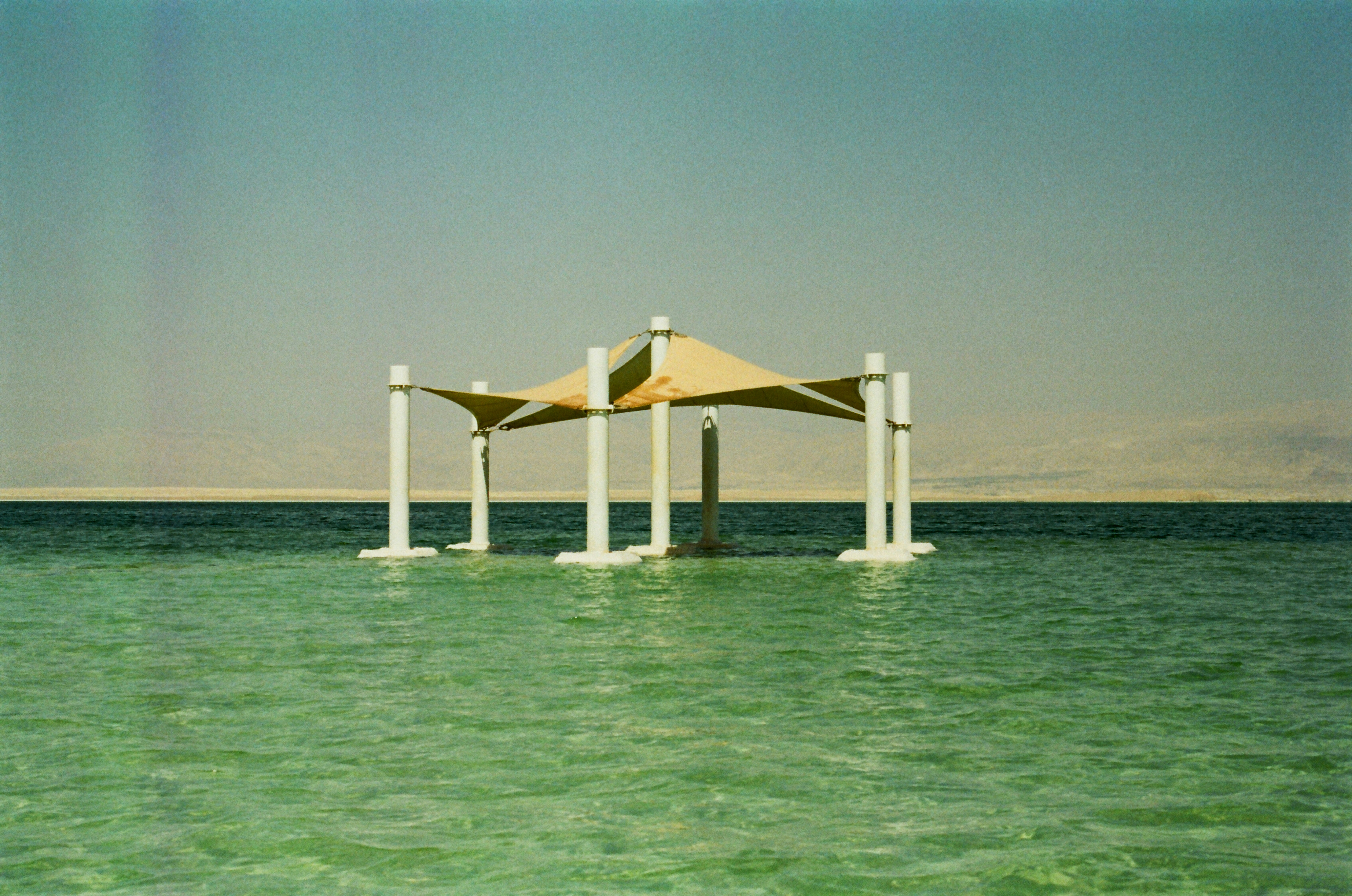 A tan fabric canopy on white pillars in the middle of turquoise water