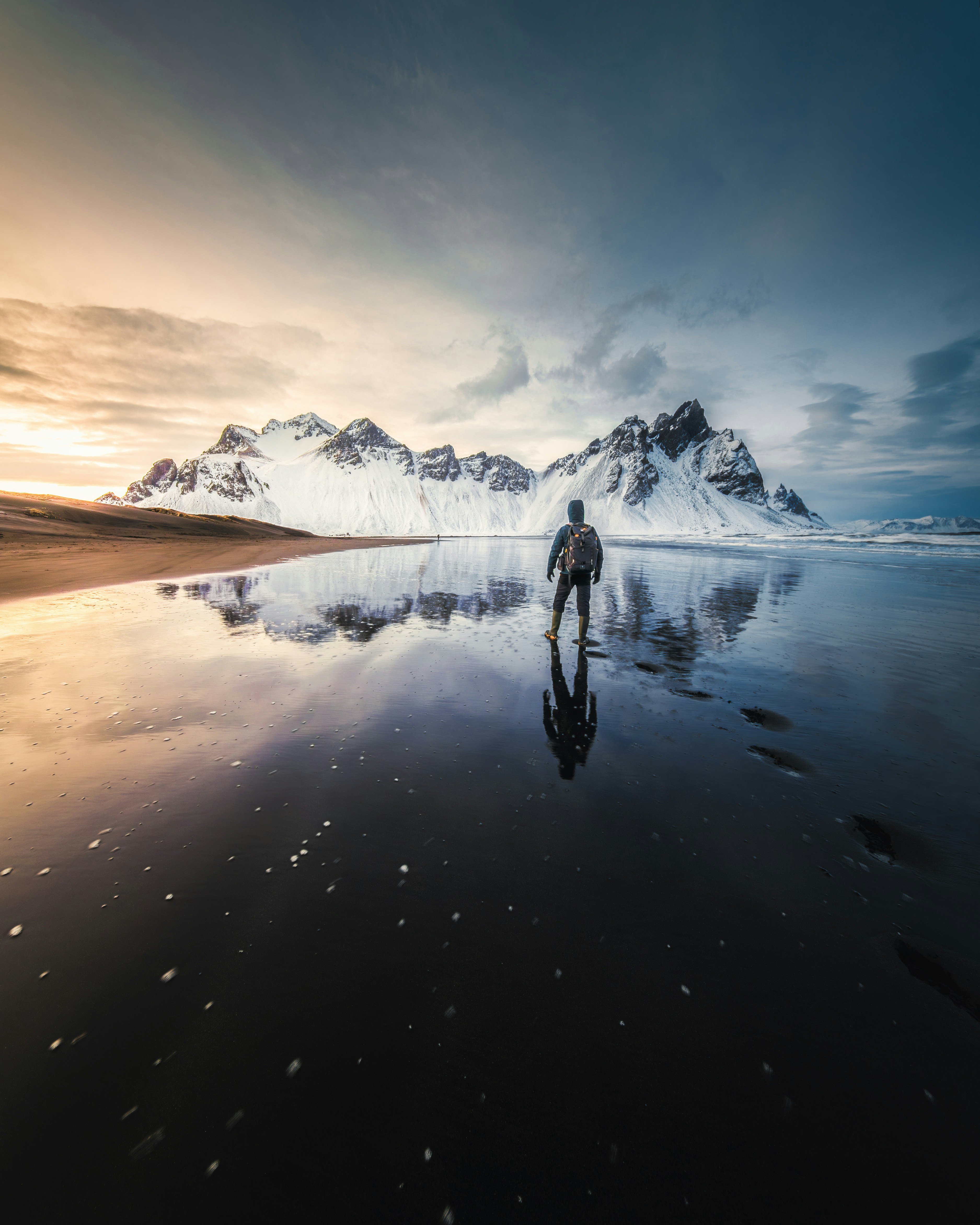 A person with a backpack on a wet black sand beach reflecting snowy mountains