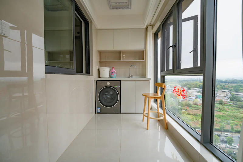 A laundry area with a washing machine, sink, and wooden stool by a window