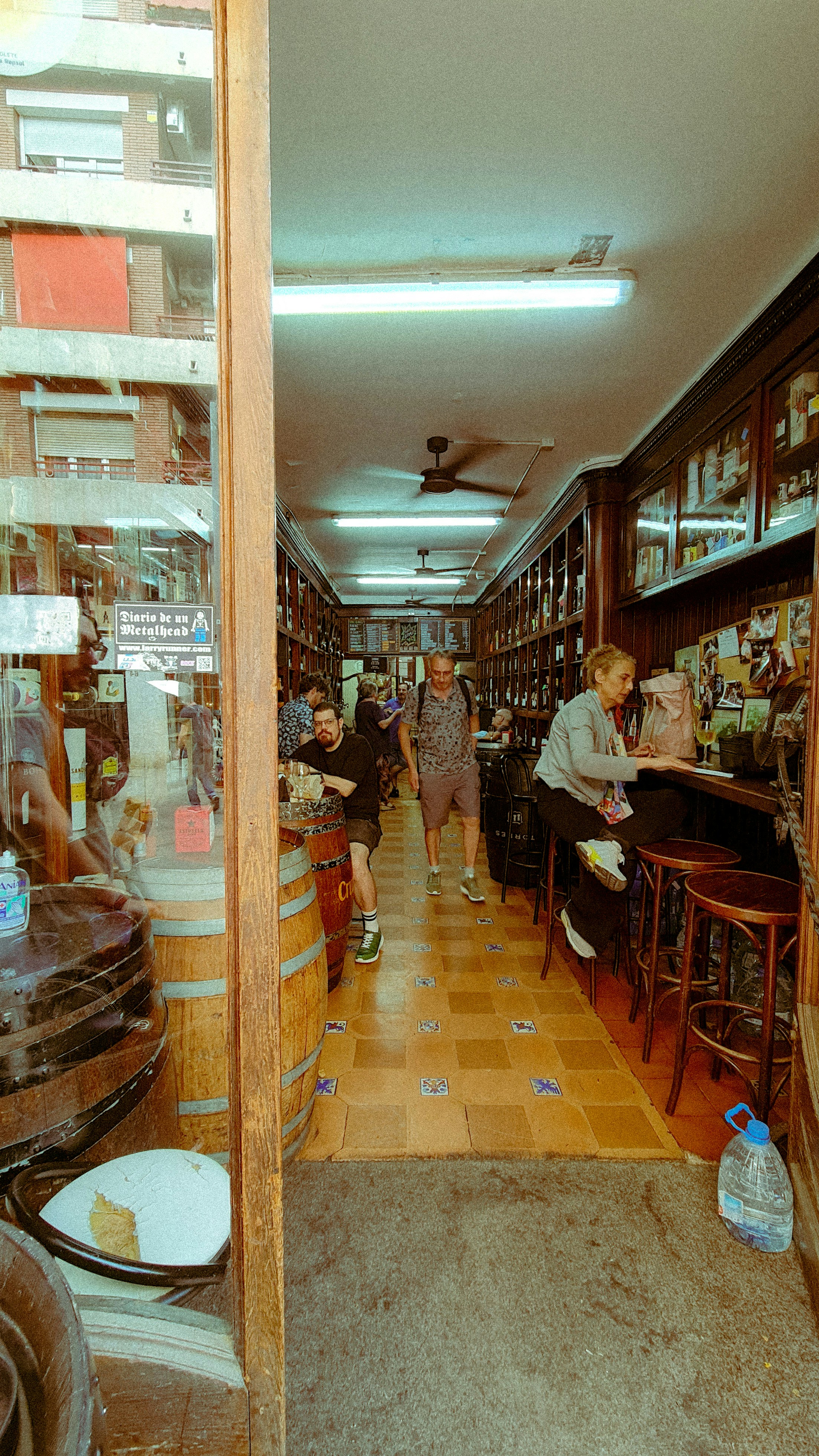 People inside a narrow bar with wooden barrels and shelves of bottles