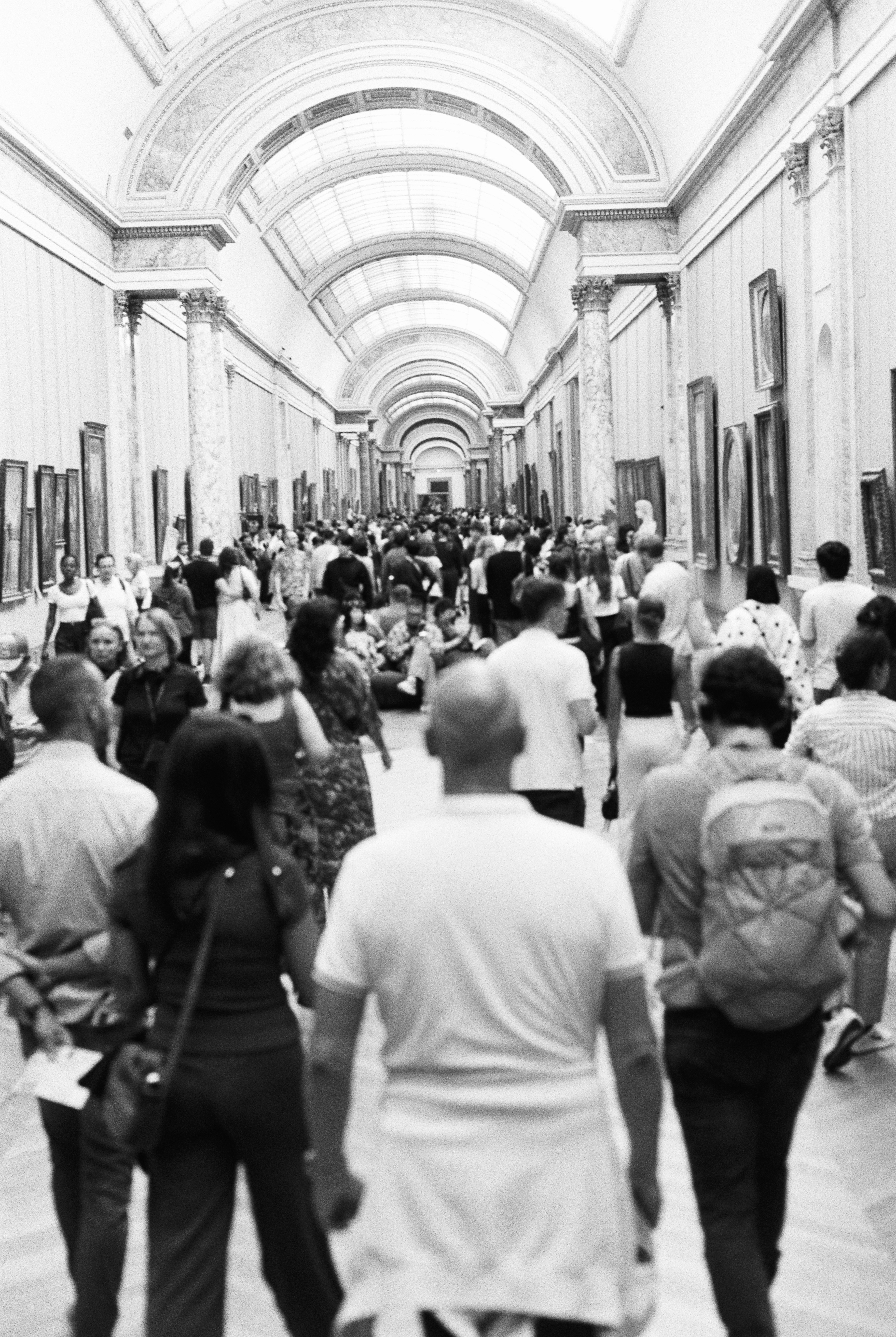 A crowd of people walking through a long, arched gallery filled with paintings