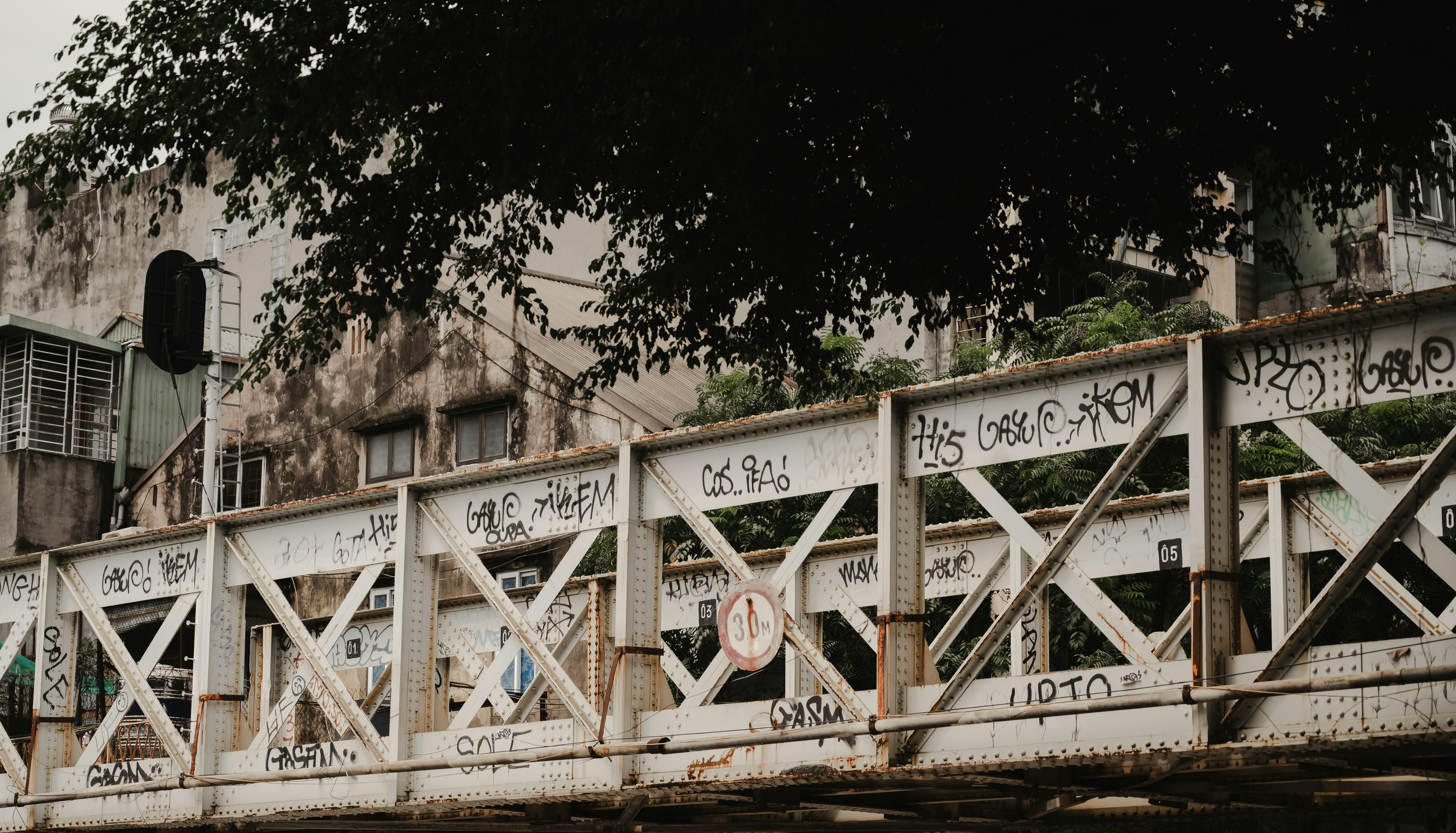 A white metal truss bridge covered in graffiti in front of old buildings