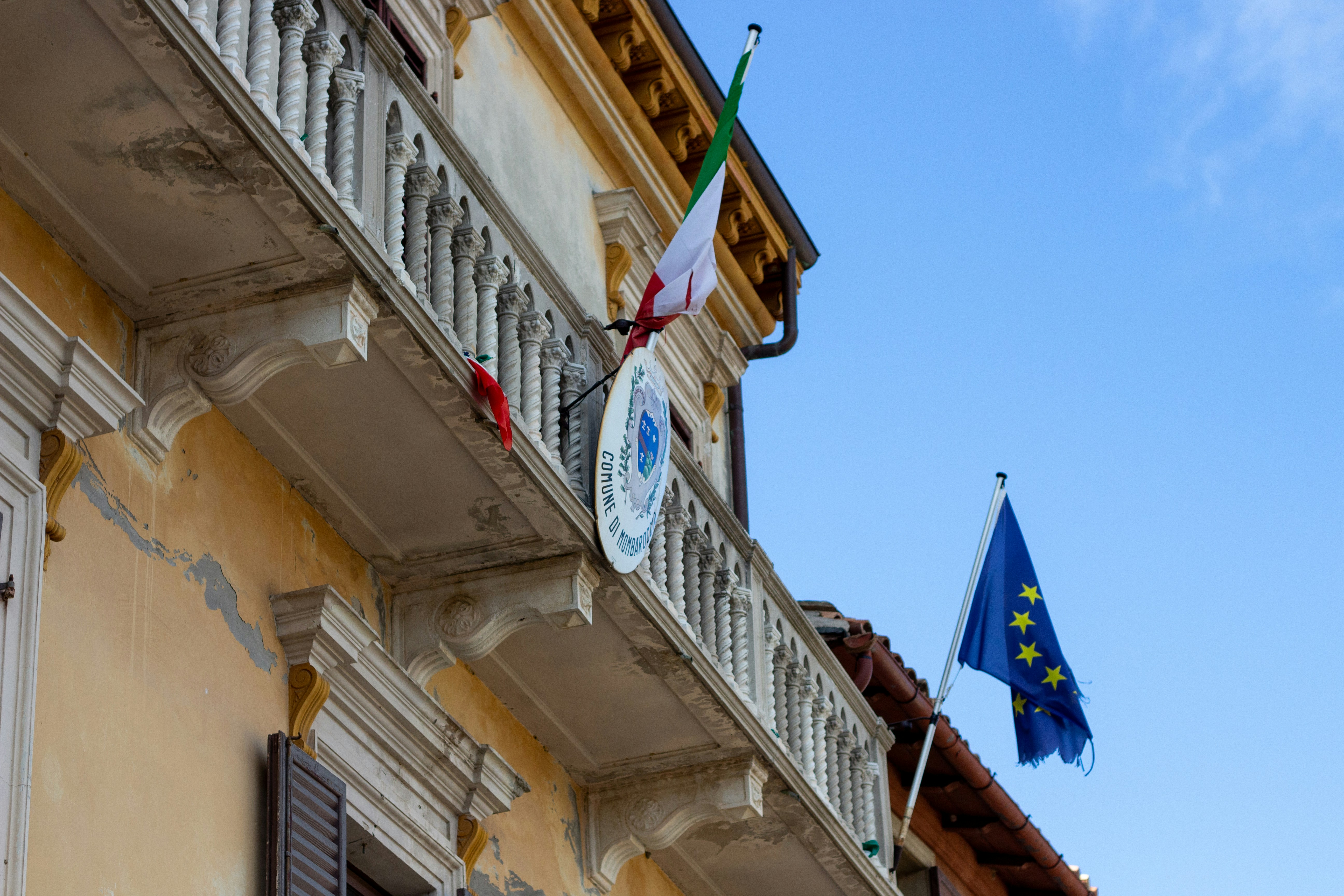 Italian and European Union flags flying from a yellow building with a stone balcony