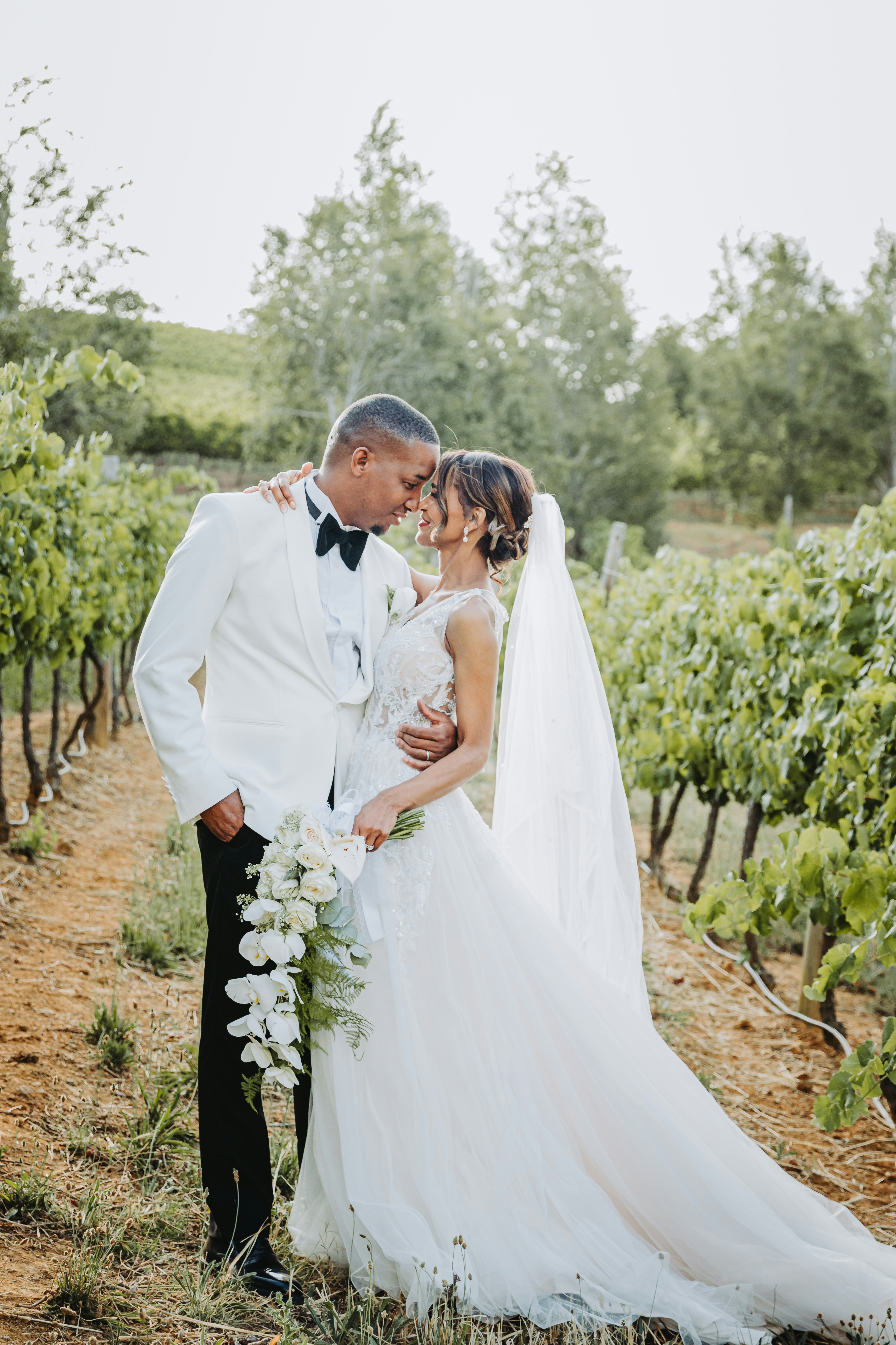 A groom in a white jacket holding a bride among green vines