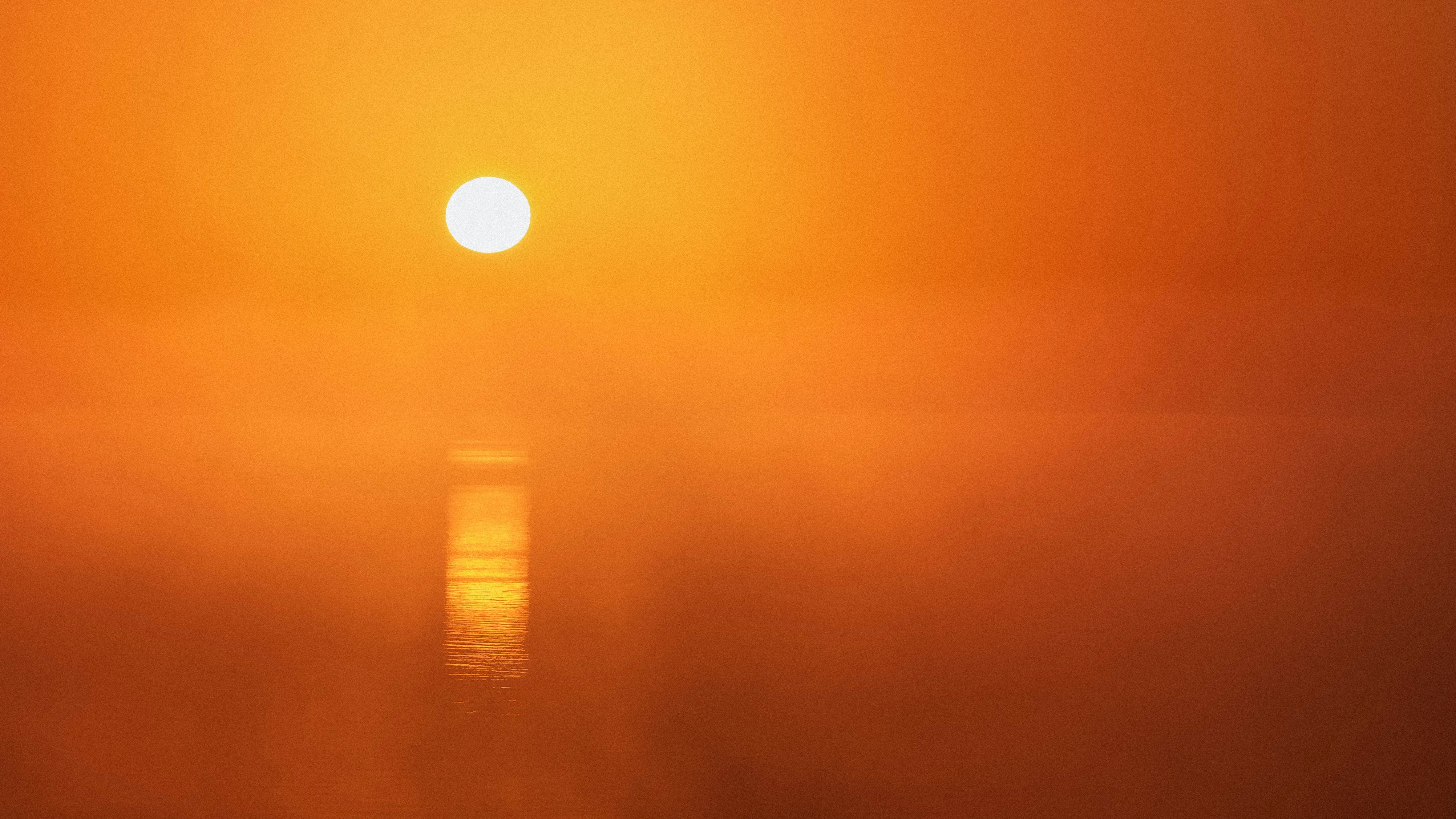 A bright sun in a hazy orange sky reflecting on calm water