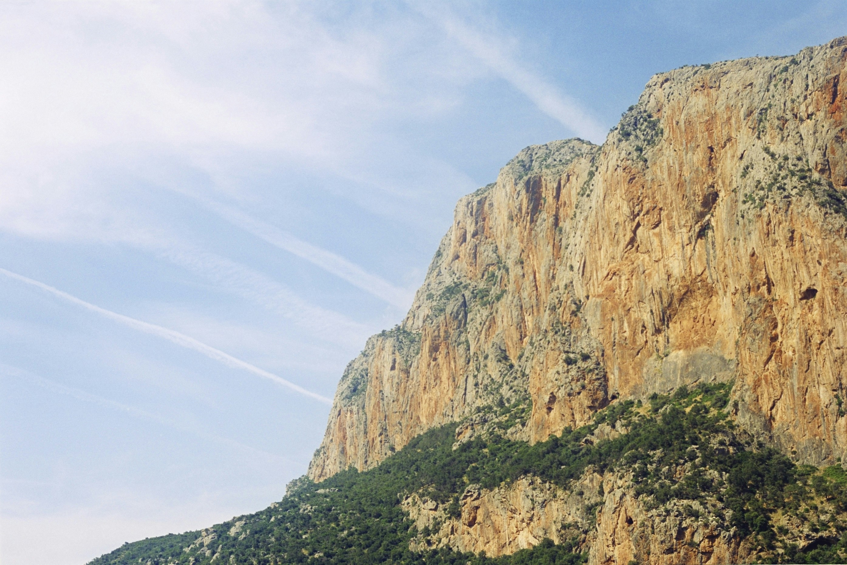 A steep orange cliff face with green trees against a blue sky with contrails